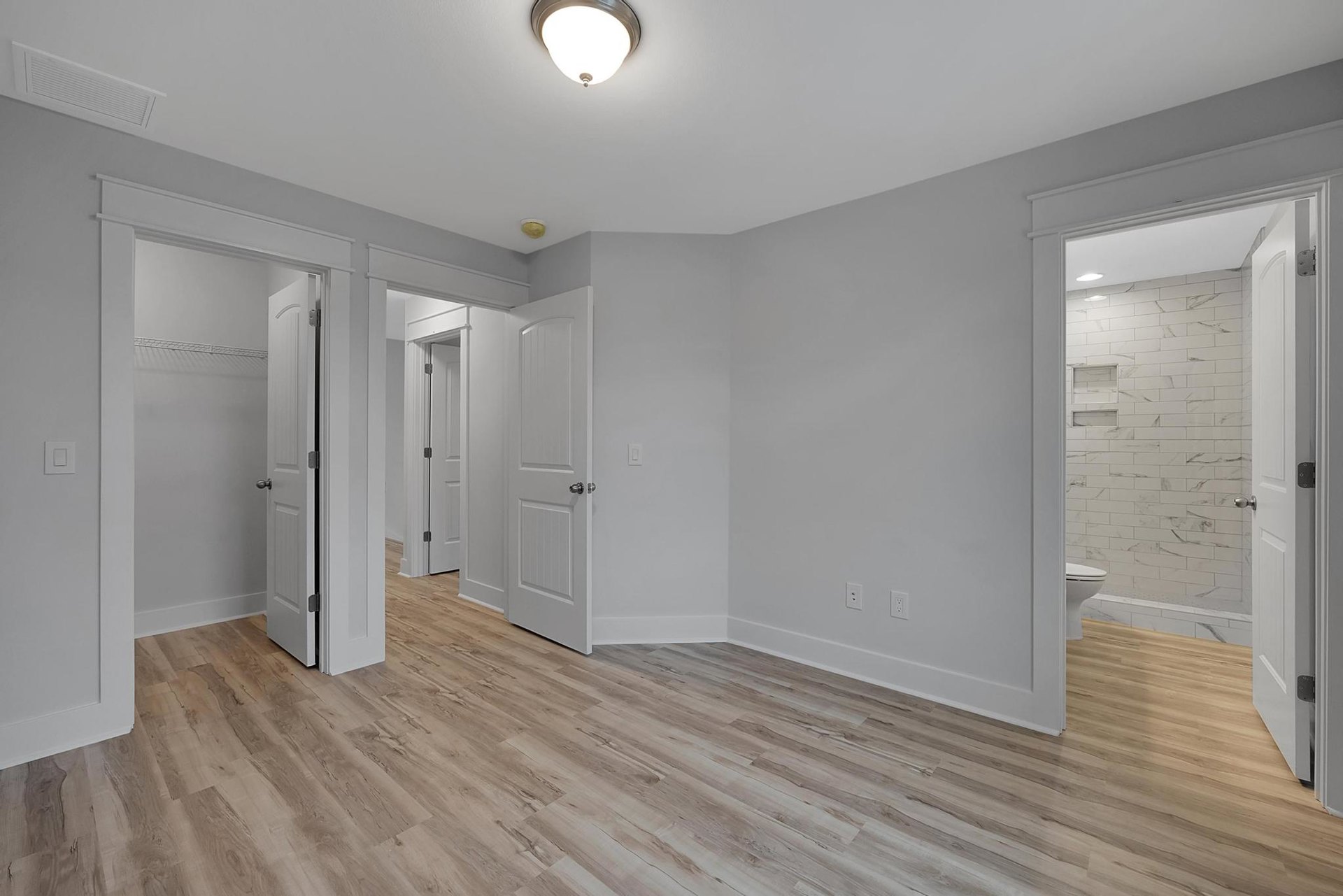 White walls, wood flooring, ceiling light fixture, white door with silver doorknob, white shelf against white tile wall