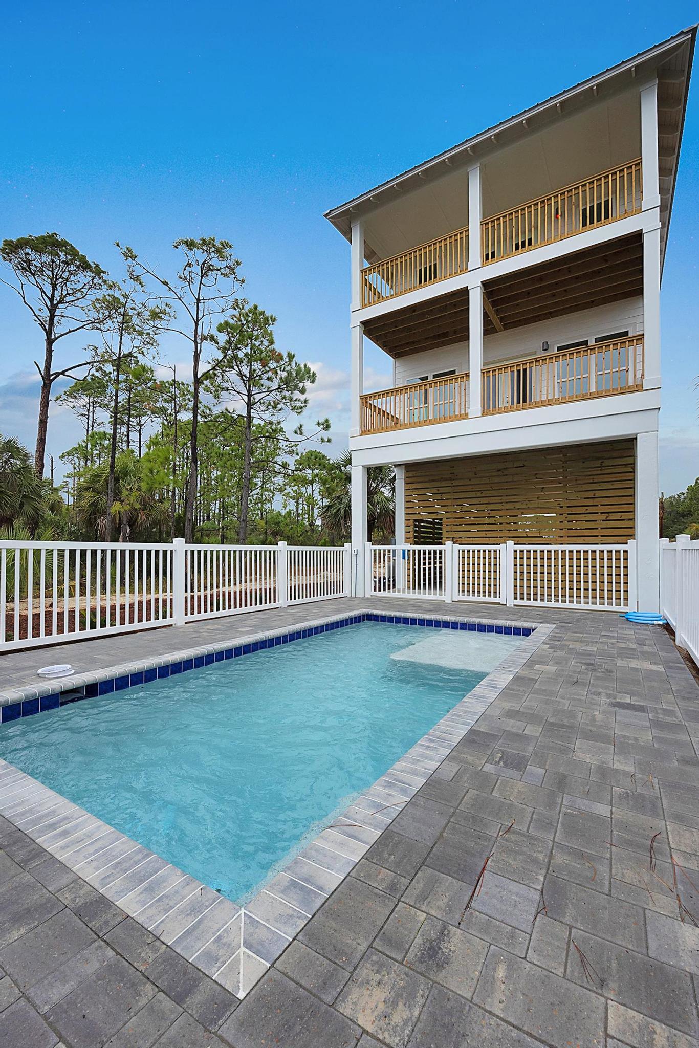 Rectangular swimming pool bordered by a white railing, adjacent to a modern building with wood siding and garage, surrounded by trees and landscaped outdoor space.