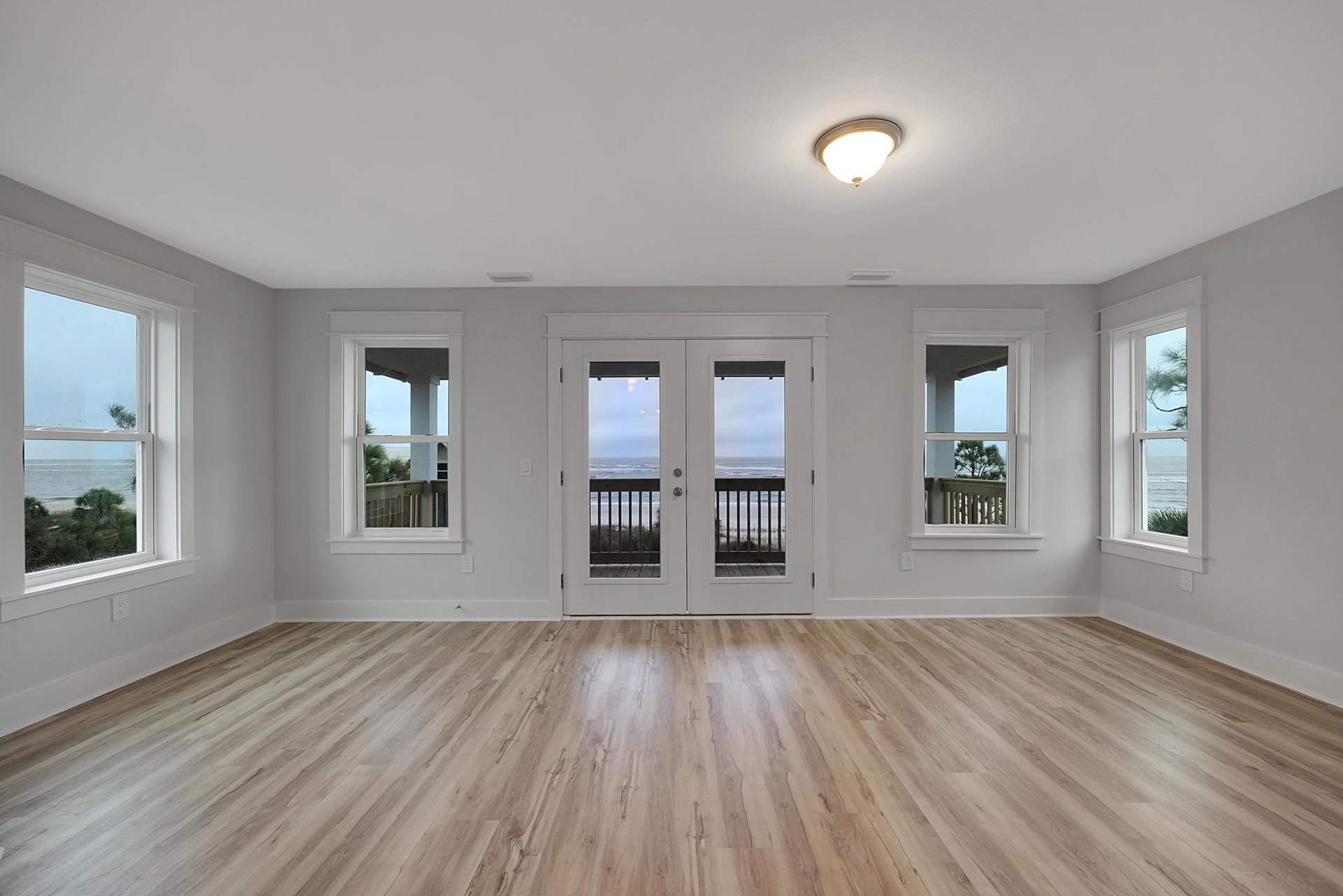 White walls and wood flooring in a bright room, featuring a white cabinet, double glass-paneled doors, ceiling light fixture, and windows with a view of a neighboring house