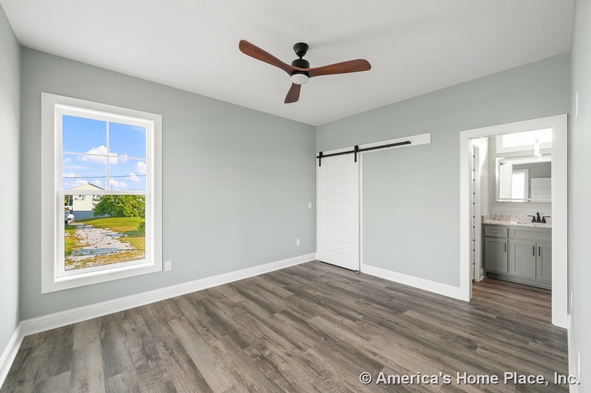Bedroom with wood-look plank flooring, large window framed in white trim, modern ceiling fan, and sliding barn door opening to en suite bathroom featuring granite countertop vanity
