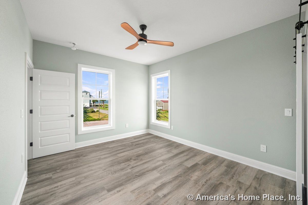 Bedroom with two large windows, light gray painted walls, wood-look vinyl plank flooring, wide white baseboards, modern ceiling fan, and white paneled door.