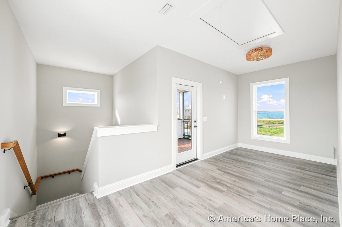 Bright upstairs landing with light gray wood-look flooring, white baseboards and trim, glass door opening to balcony, large window offering ocean view, wood handrail staircase