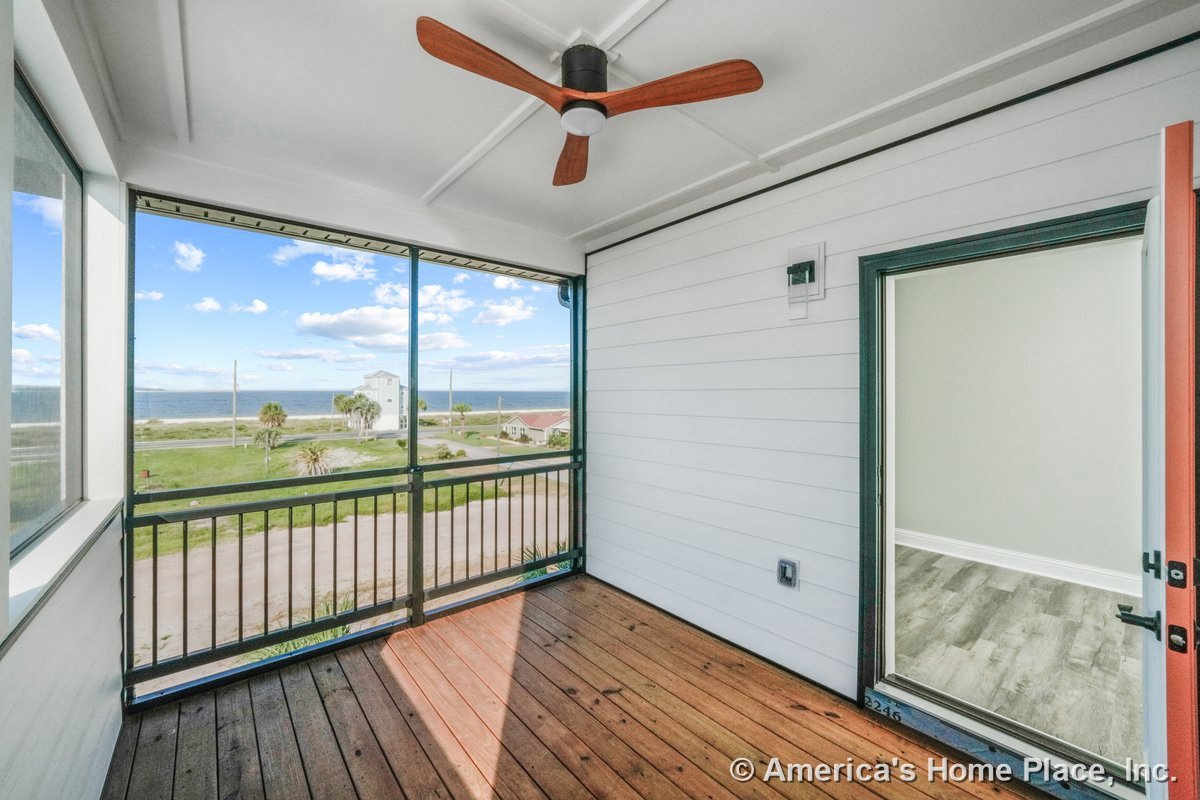 Screened porch with wood plank flooring, horizontal wall siding, modern ceiling fan, metal railing, and glass door with decorative trim; built-in lighting and coastal view visible.
