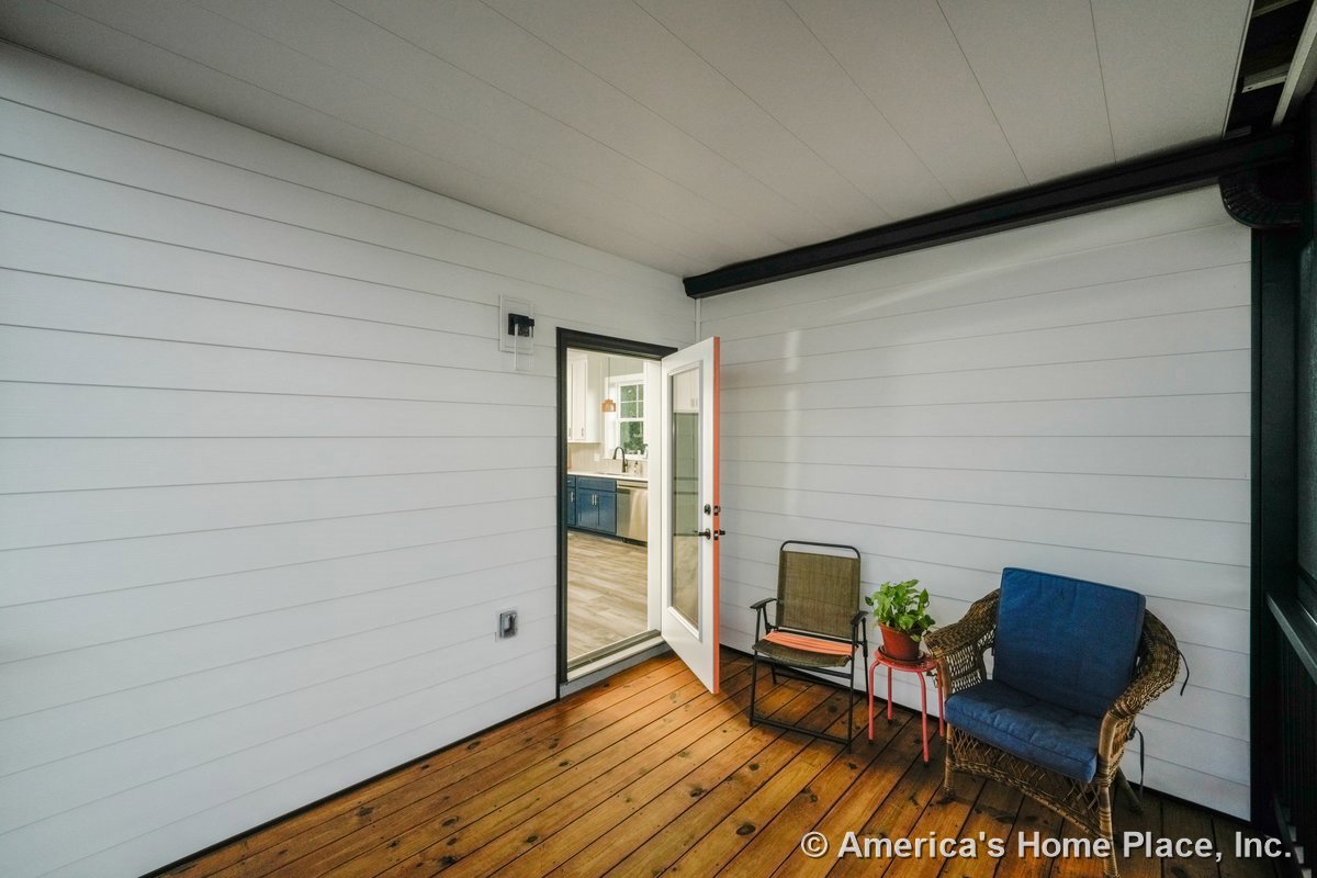 Screened porch with horizontal white siding, wood plank flooring, glass door entry, built-in wall light fixture, and interior view into kitchen.