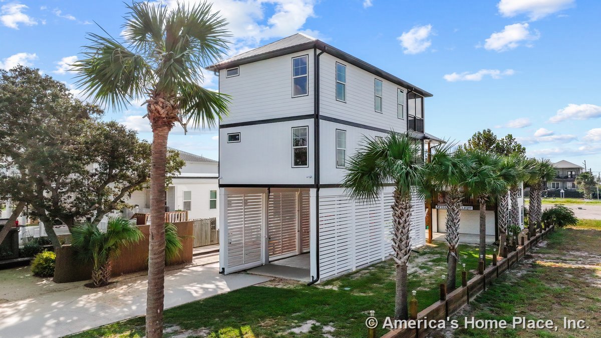 Three-story coastal home with white horizontal siding, black exterior trim, ground-level carport enclosed by slatted panels, multiple rectangular windows, and paved driveway.