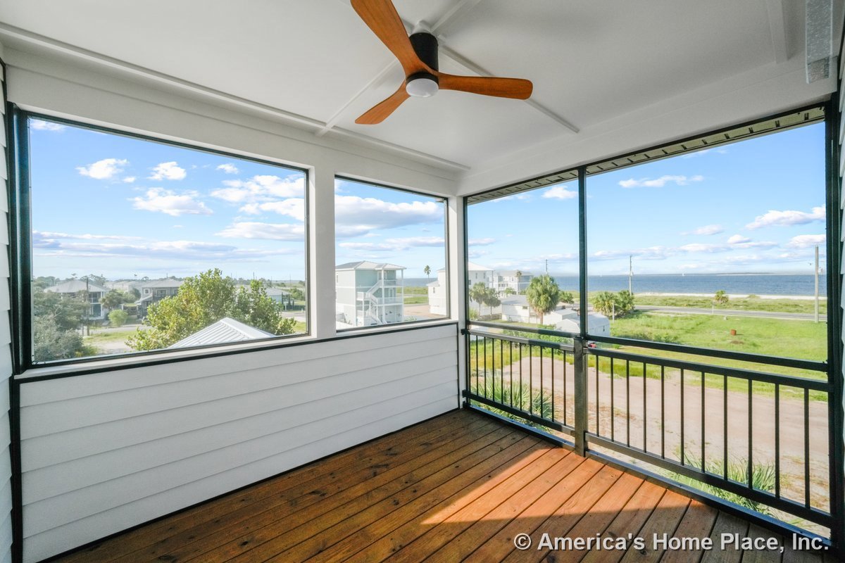Screened porch with wood plank flooring, white horizontal siding, black metal railing, ceiling fan with wood blades, and large windows overlooking the ocean.