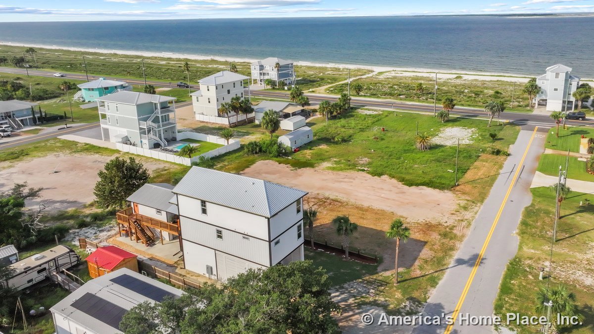 Elevated coastal home with metal roofing, raised foundation, exterior staircase, multiple stories, large windows, and siding overlooking driveway, yard, and beach view.