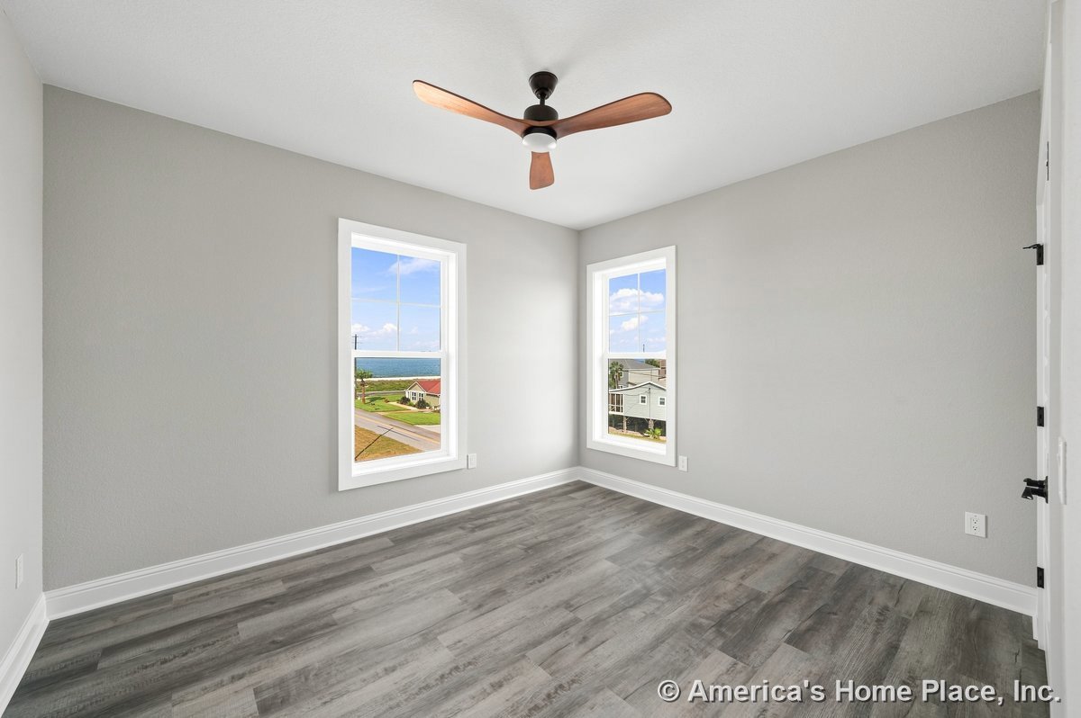 Bedroom with two large windows framed in white trim, gray painted walls, wood-look vinyl plank flooring, ceiling fan with wood blades, and baseboard molding, filled with natural