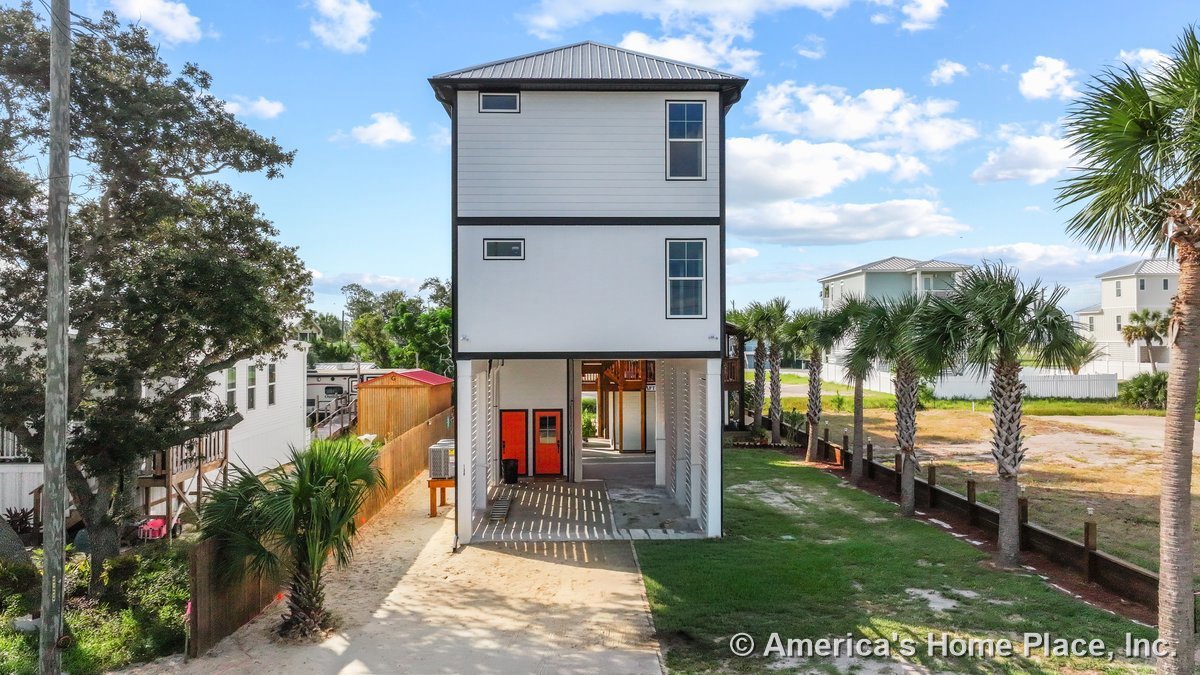 Elevated coastal home with metal roof, multiple rectangular windows, vertical siding, covered parking area beneath, and red entry door.