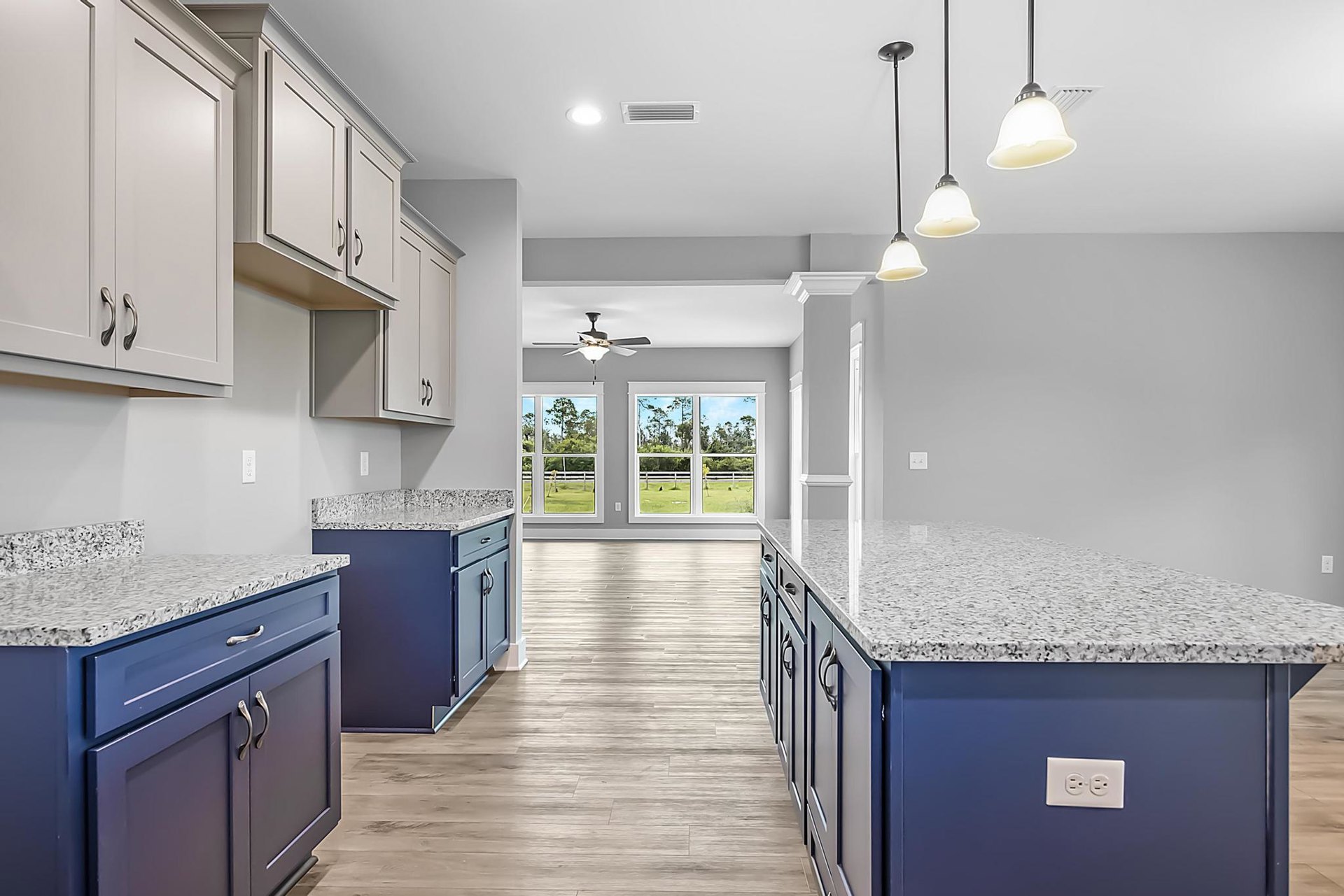 Blue kitchen cabinets with white marble countertops, stainless steel sink, ceiling fan with light fixture, electrical outlet on backsplash, windows showing trees and fence outside
