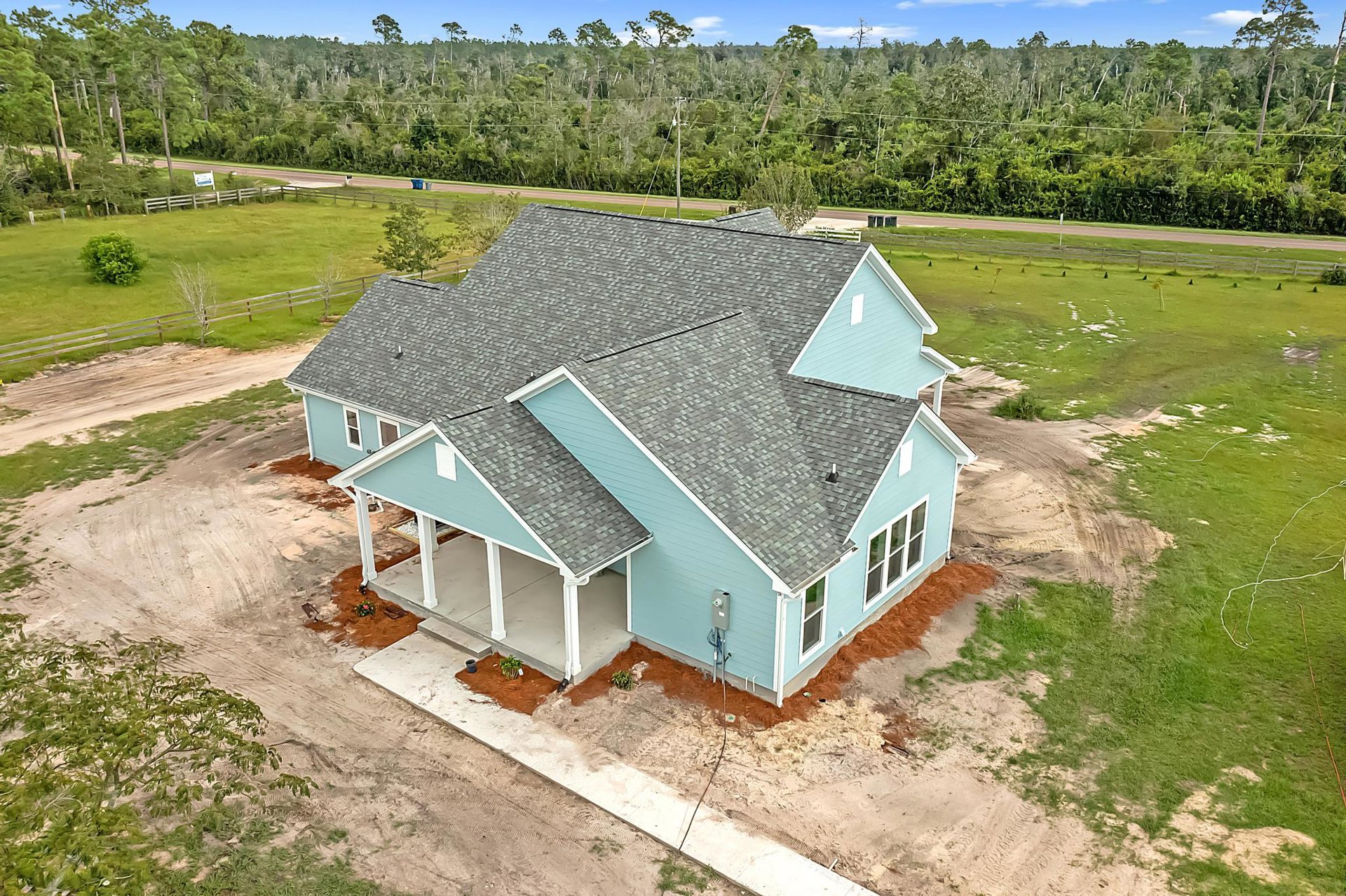 Blue cottage-style home with white trim, covered front porch, fenced yard, manicured lawn, mature trees, and overhead power lines against a clear sky.