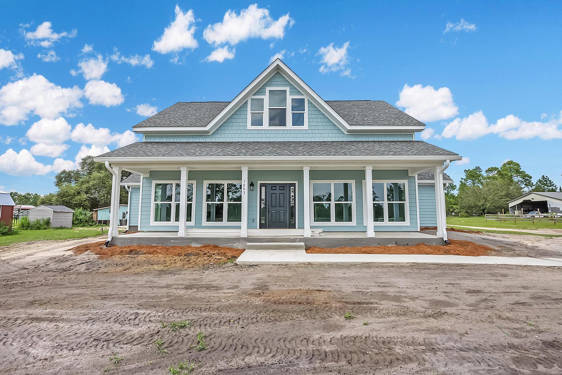 Two-story home with white siding, black door with glass panes, white-framed windows, covered porch, dirt driveway, and surrounding trees under a cloudy sky