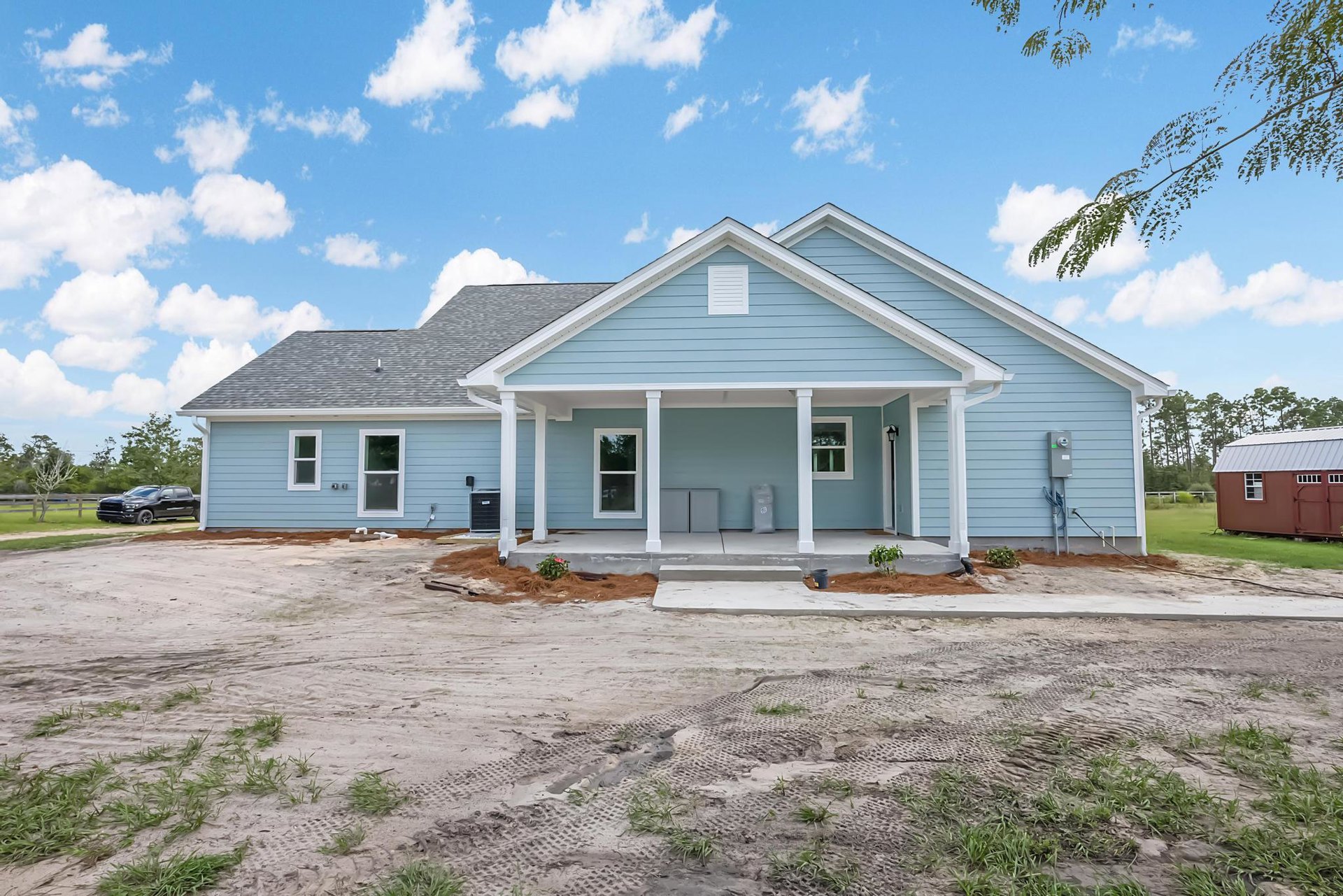 Blue house with white trim and white framed windows, red shed with white roof nearby, black car driving on dirt road, white door with vertical lines, trees and cloudy sky in