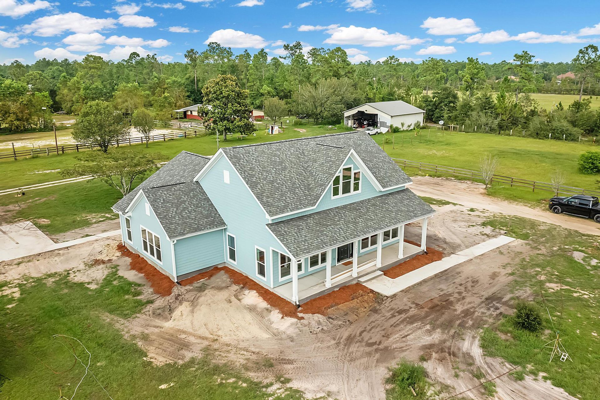 Blue house with white trim surrounded by mature trees, patchy grass, and exposed dirt; black car parked on unpaved driveway under partly cloudy sky.