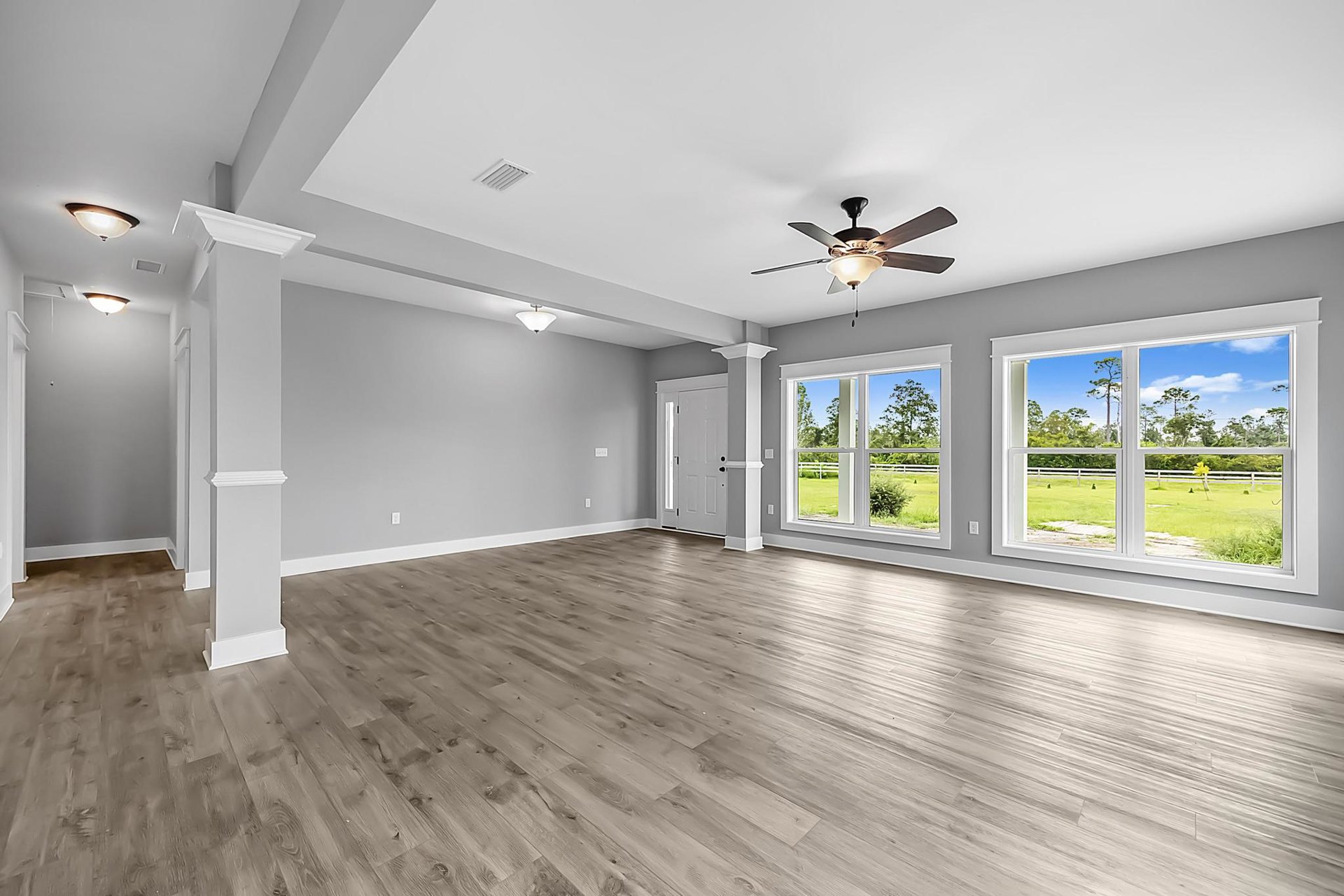 Ceiling fan with light fixture, white vent on ceiling, wood laminate flooring, white paneled door, large windows overlooking grassy field, neutral walls