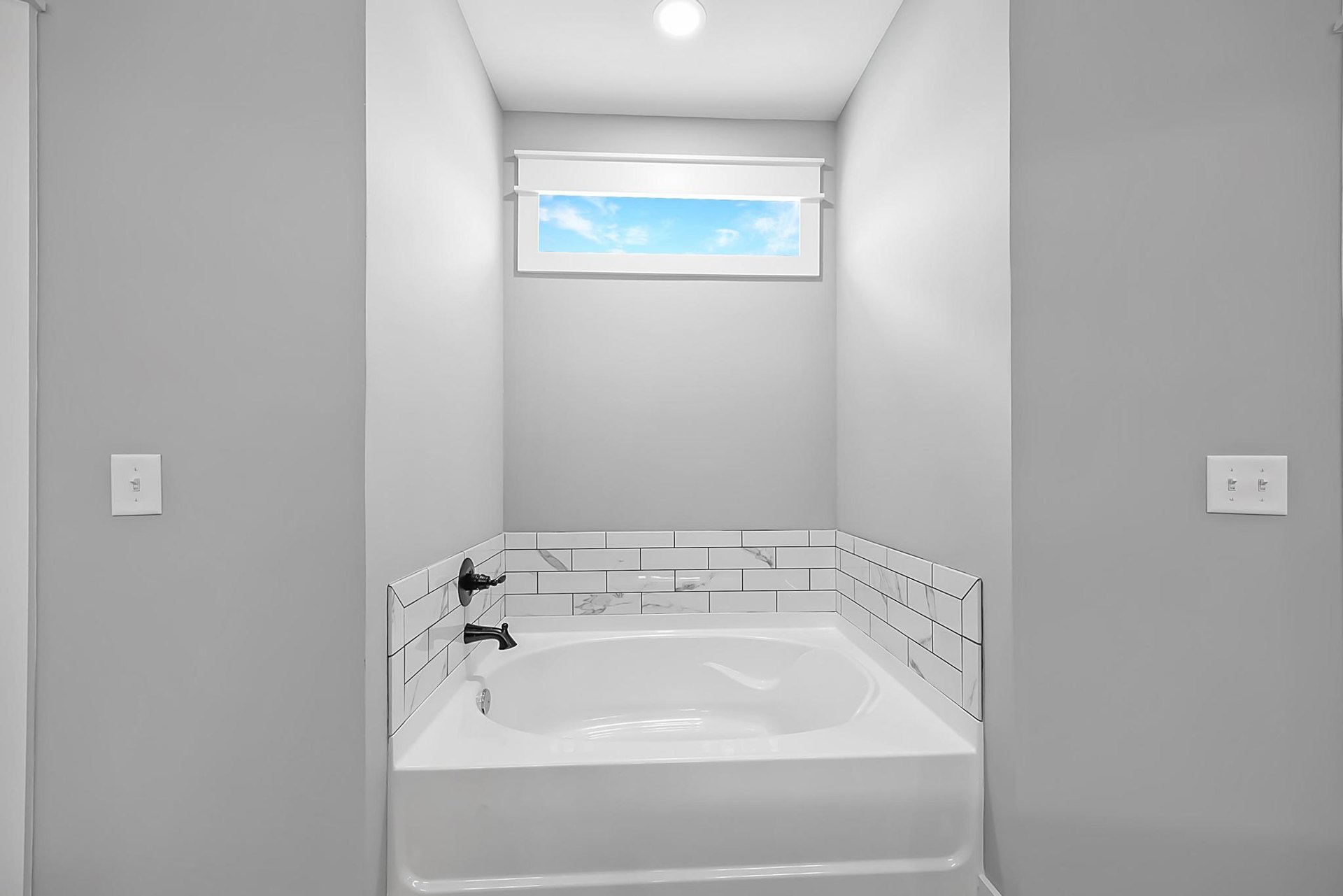 Freestanding white bathtub with black faucet against tiled wall, blue sky and clouds visible through window, double white light switch on adjacent wall