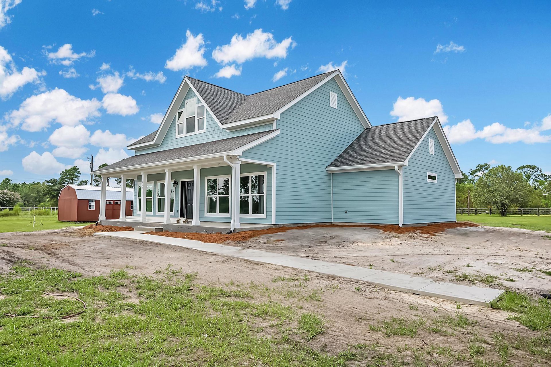 Blue house with white trim and blue roof, driveway bordered by grass, leafy tree nearby, red shed with windows in background, cloudy sky overhead.