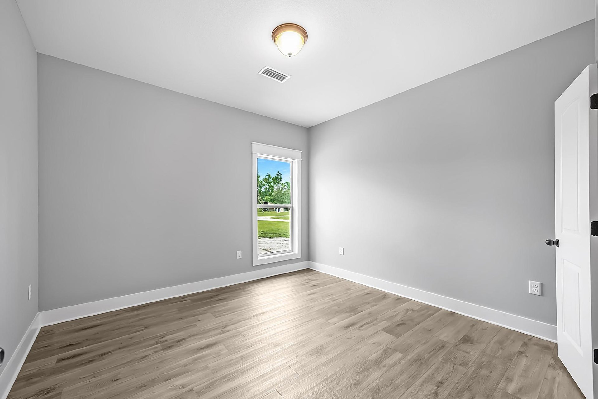 Sunlit room featuring wide-plank hardwood flooring, white walls, a large window overlooking a green lawn, recessed ceiling vent, modern ceiling light, and a white paneled door.