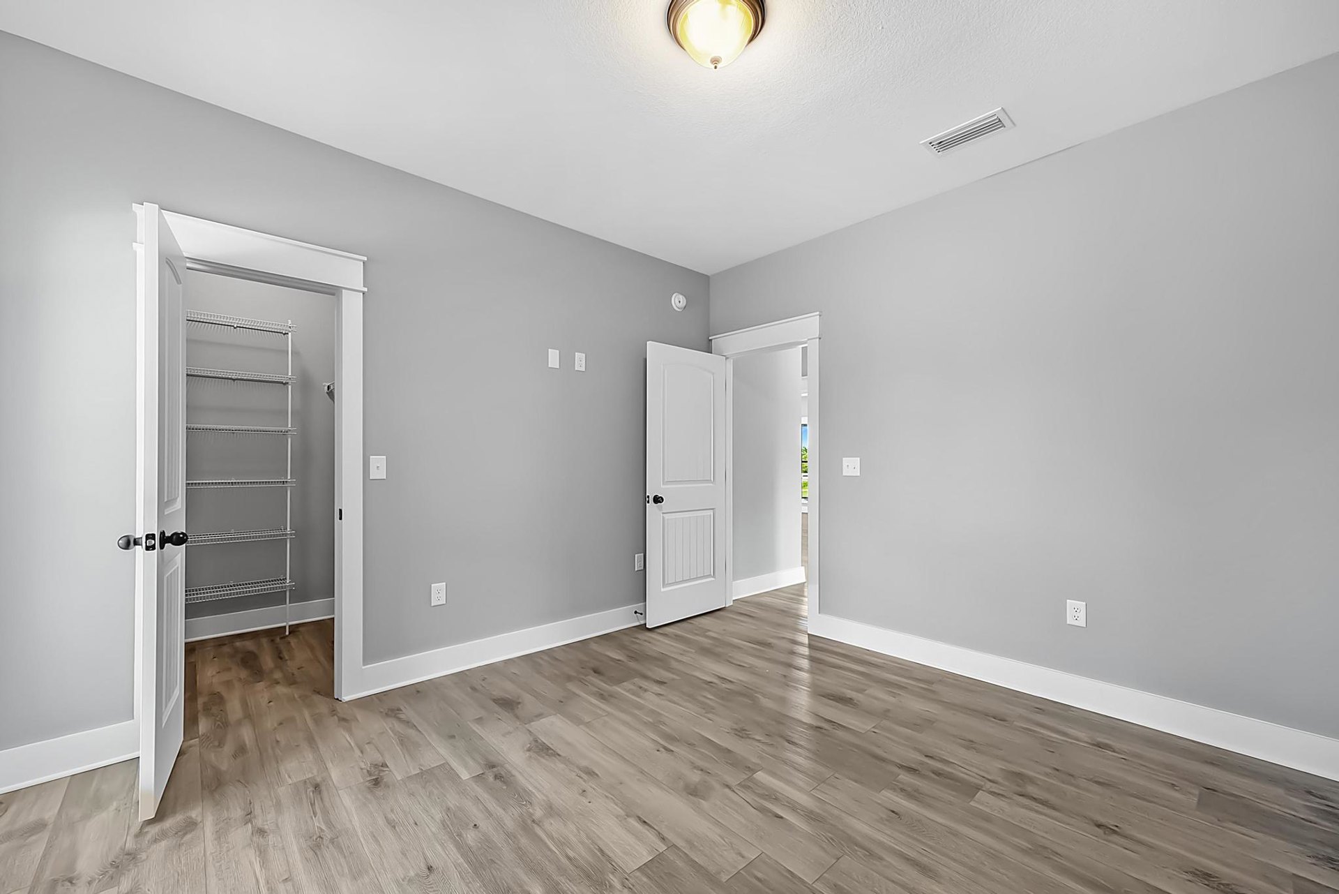 White walls and door with black hardware, wood flooring, built-in white closet with metal rods, open shelving, and ceiling light fixture.