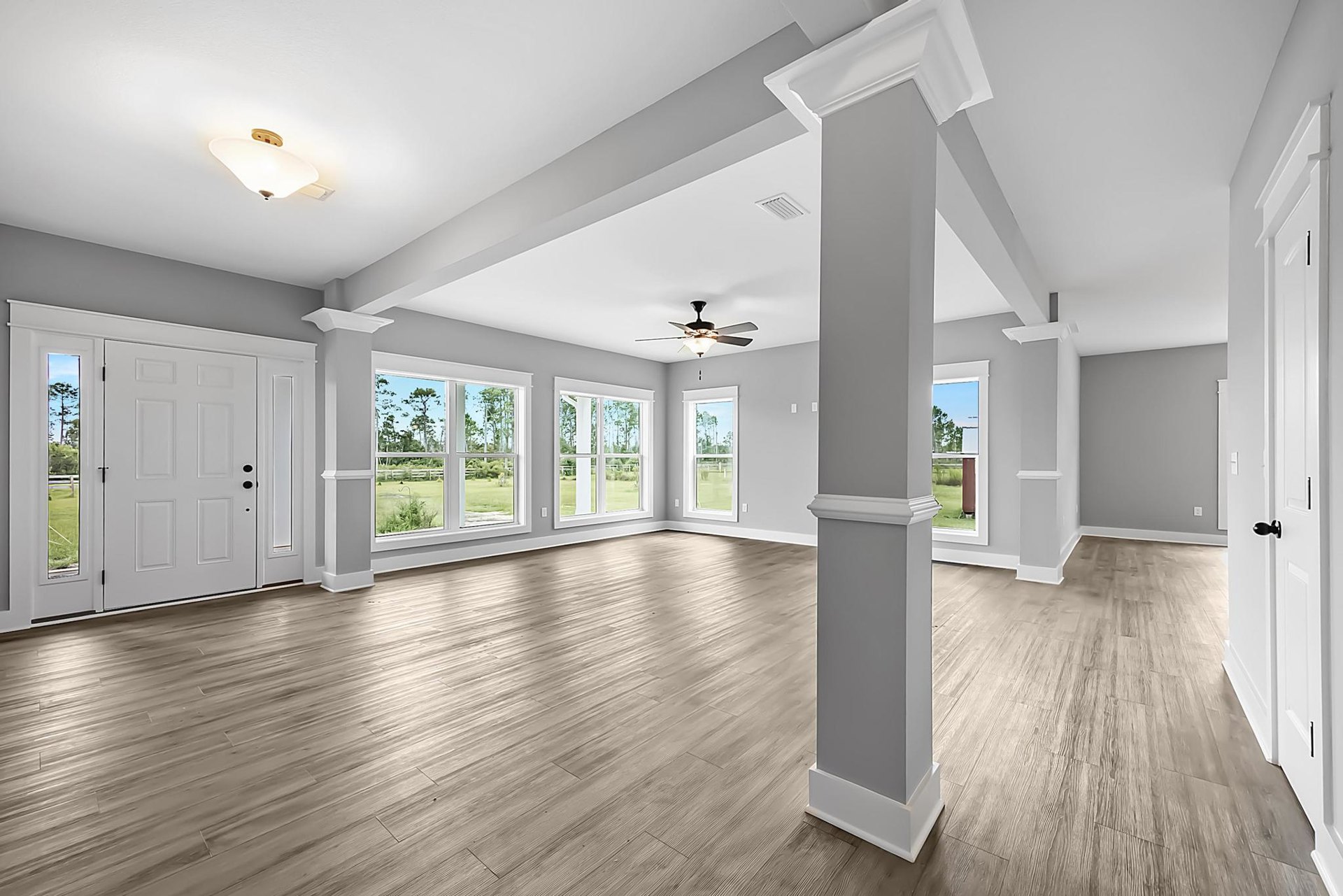 Spacious room featuring white columns, wood flooring, ceiling fan with light, multiple windows overlooking a lawn, and a white door with glass panels