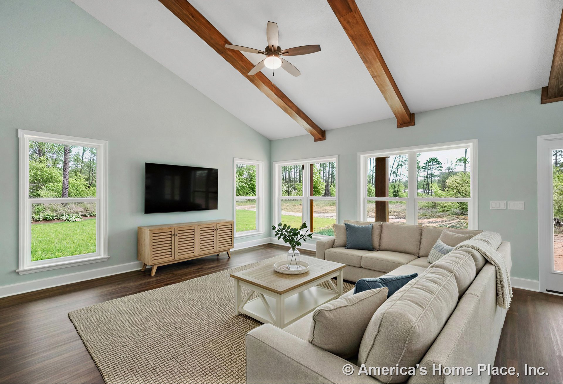 Vaulted ceiling with exposed wood beams, wall of large windows with white trim, wood-look flooring, ceiling fan with light, wall-mounted TV, and glass exterior door.