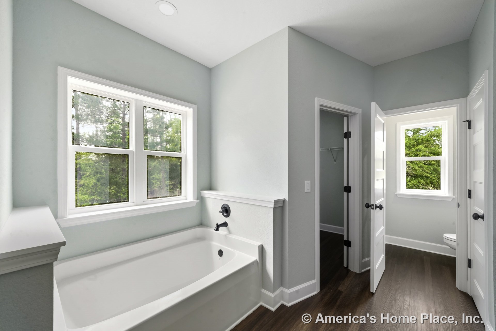 Built-in soaking tub with wall-mounted faucet positioned beneath a double window with white trim, wood-look plank flooring, light blue painted walls, white paneled doors, recessed