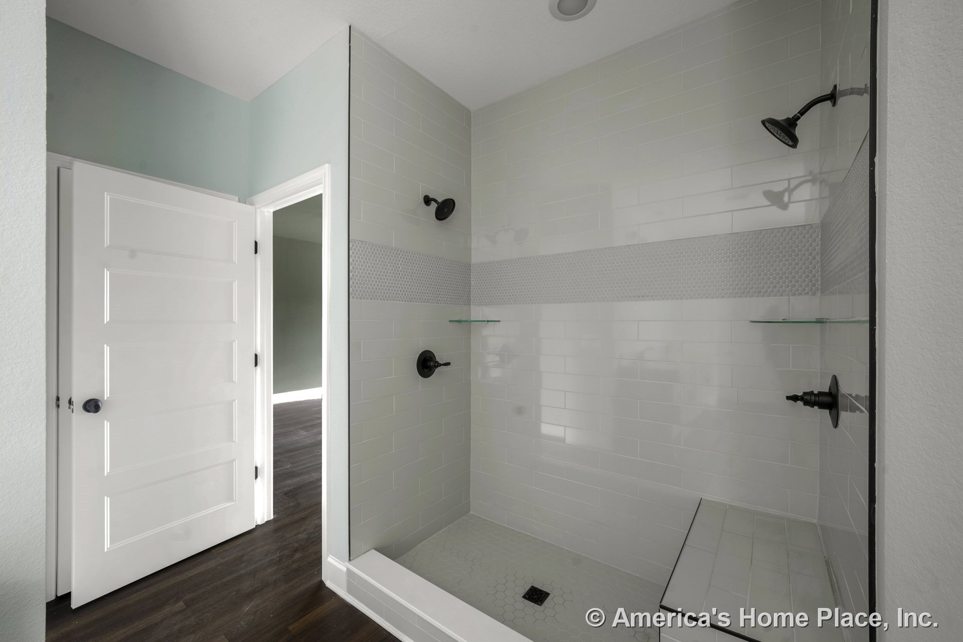 Walk-in shower with white subway tile, built-in bench, double shower heads, glass corner shelves, hexagonal floor tile, and horizontal accent tile band; white paneled door and dark