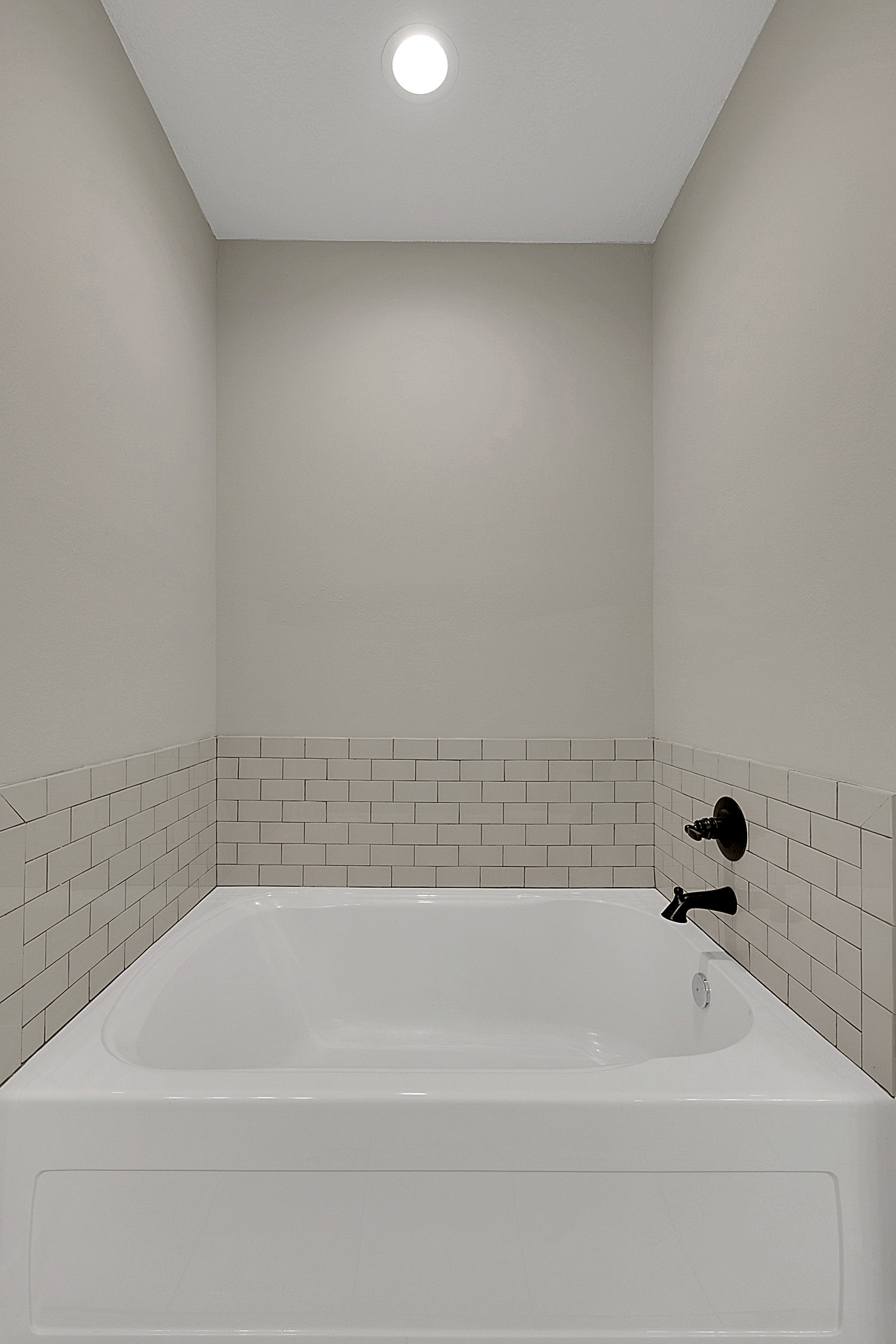 White freestanding bathtub with chrome faucet set against a white tile wall, circular ceiling light above, and white tile border detailing