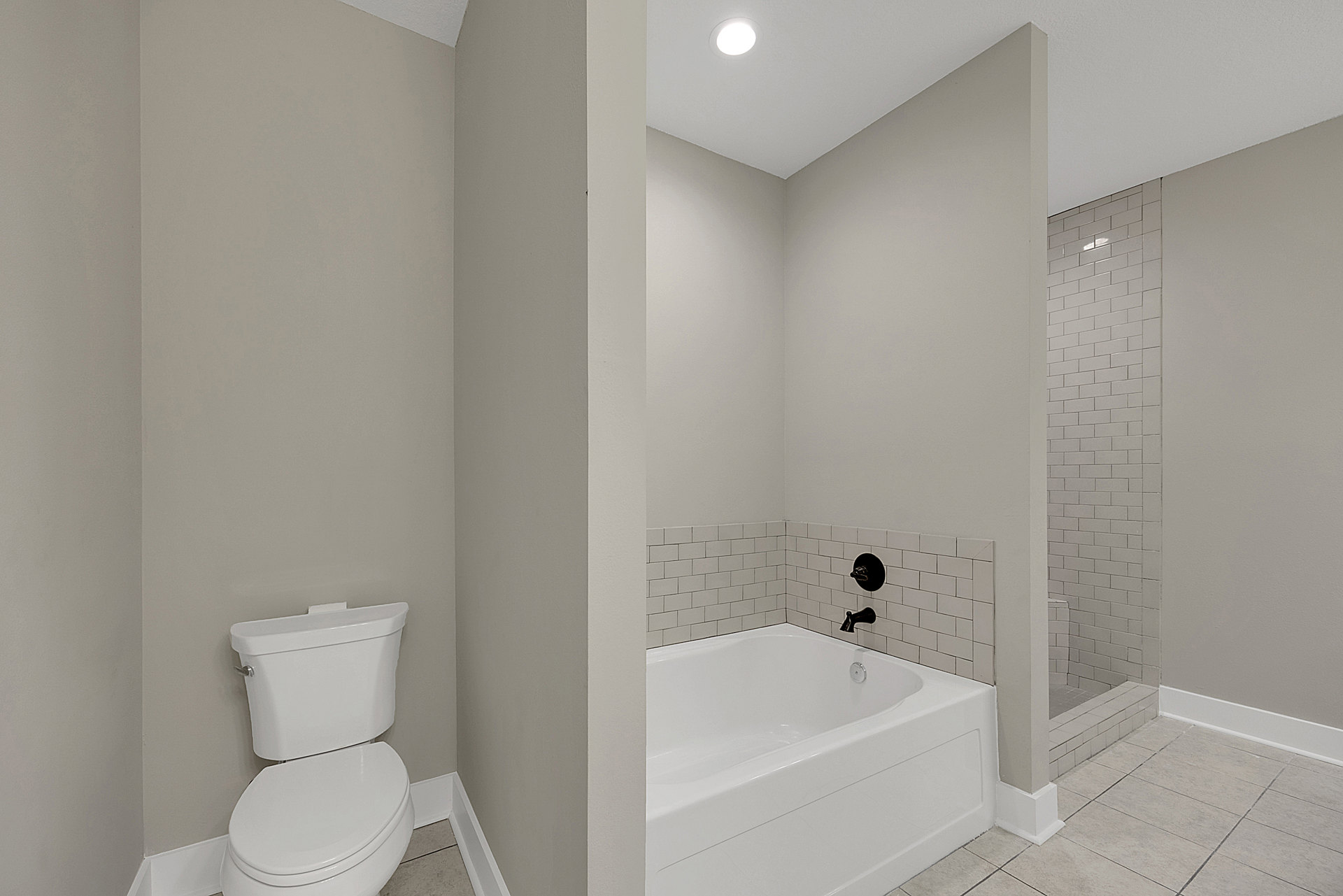 Bathroom with white bathtub featuring black faucet, glass-enclosed shower, white toilet with lid up, light fixture in ceiling, neutral tile walls and floor