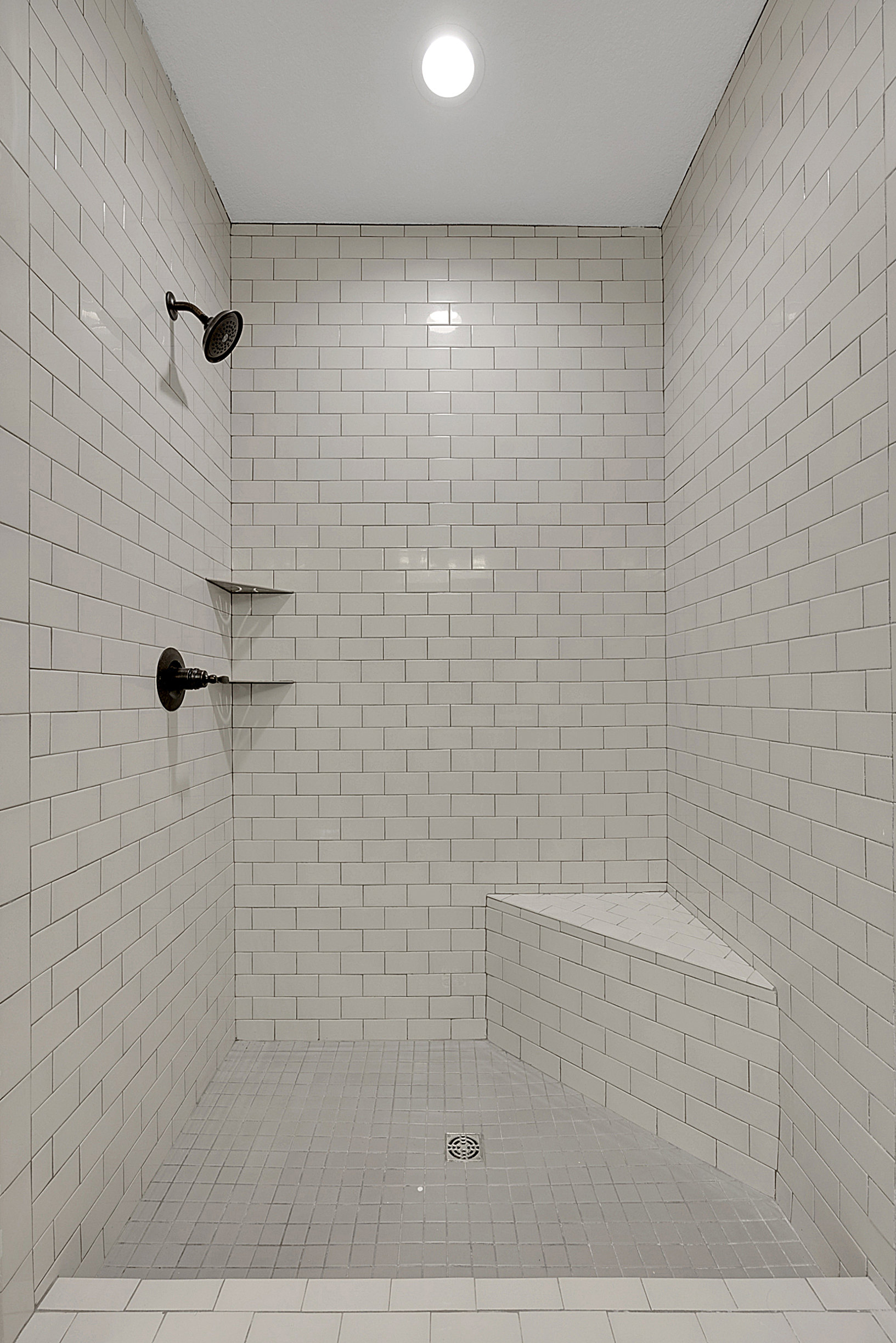 Walk-in shower with light gray tile walls, built-in bench, chrome shower head mounted on wall, and recessed ceiling light