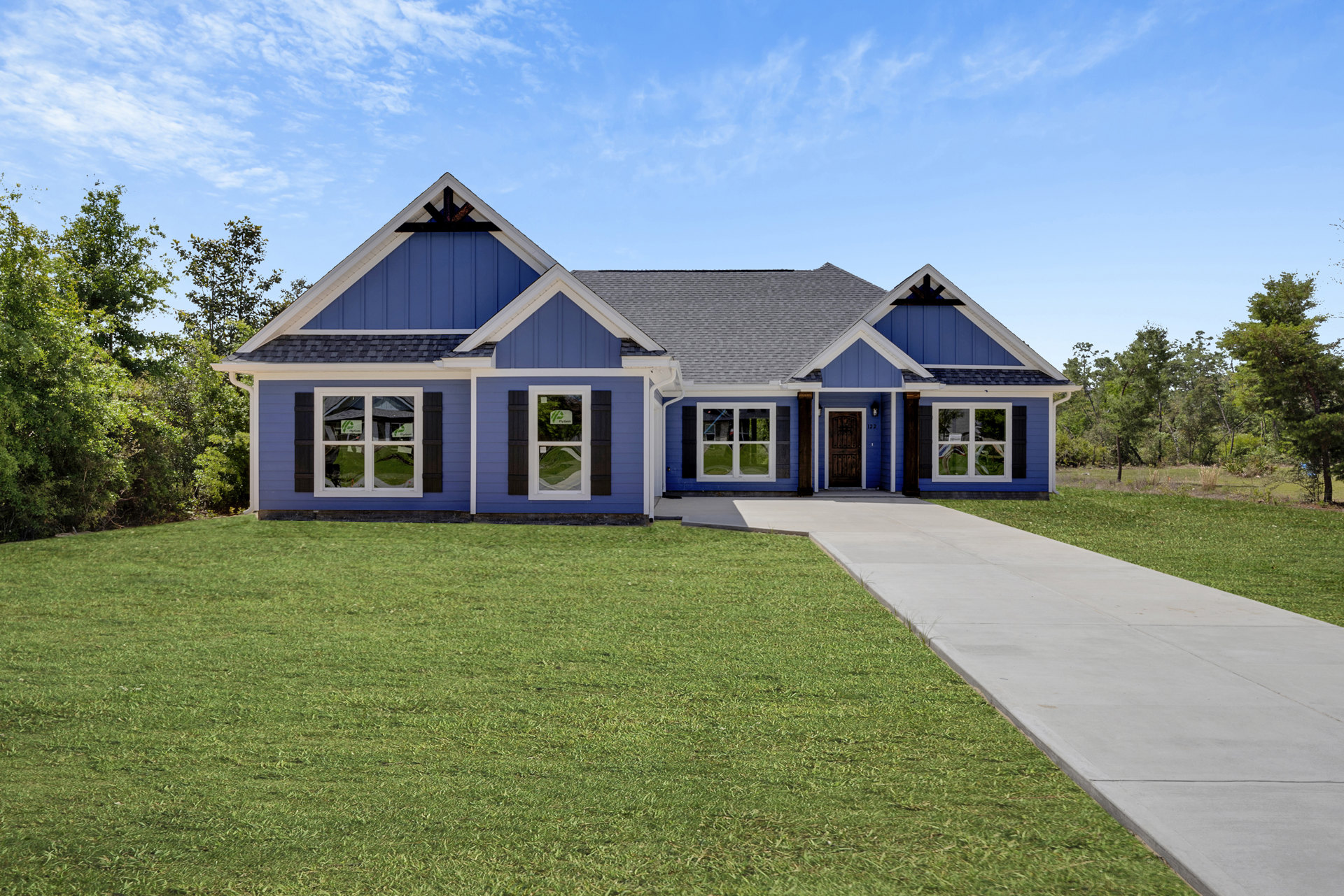 Blue house with white trim, blue front door, white-framed windows, concrete driveway, green lawn, sidewalk, and trees under a partly cloudy sky