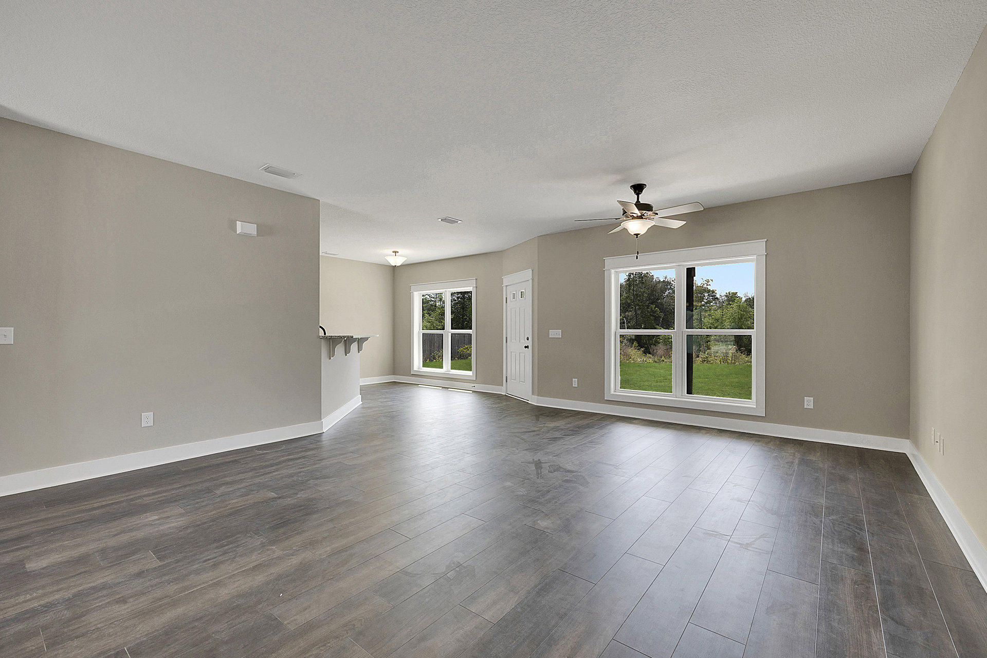 Spacious room with light wood flooring, white walls, ceiling fan with light fixture, large window overlooking backyard trees, and white door with black handle