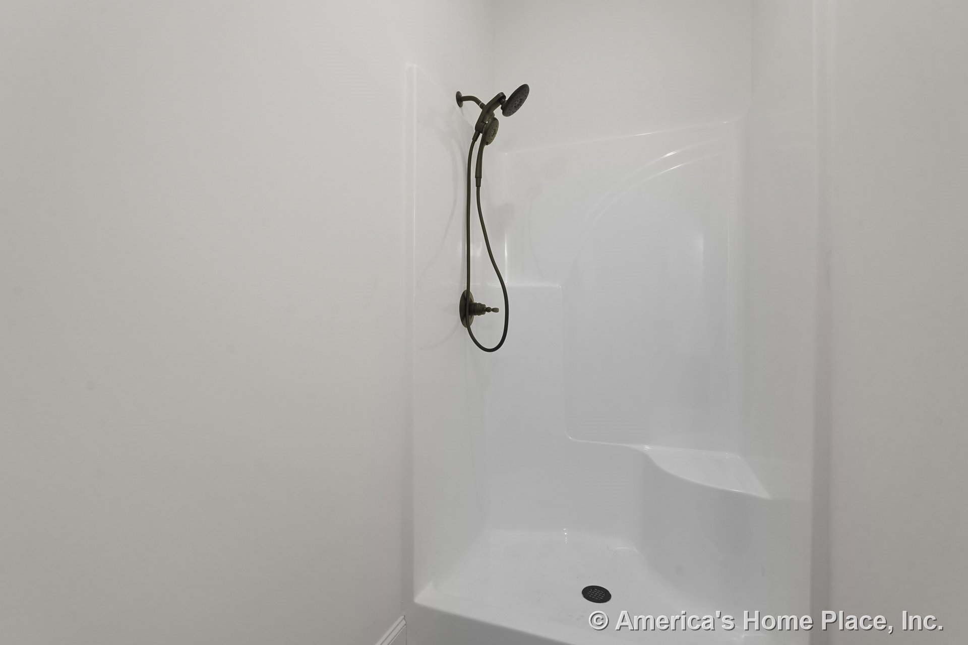White fiberglass shower enclosure with built-in seat, bronze handheld shower fixture, white wall trim, and floor drain.