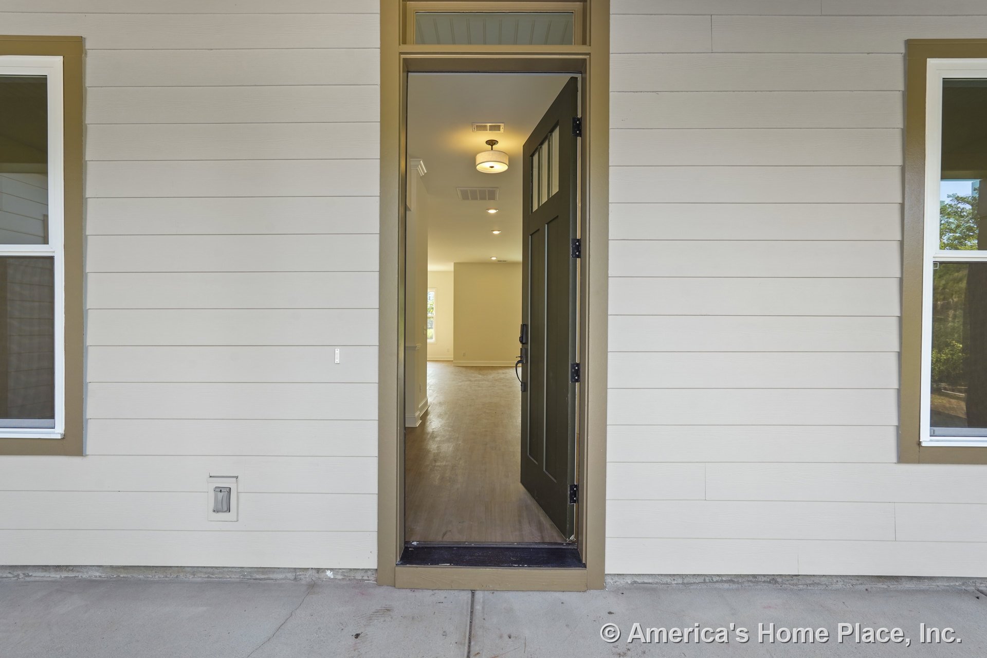 Paneled front door with transom window, brown trim, and horizontal exterior siding opens to entry hall with wood flooring, recessed lighting, and visible open interior layout