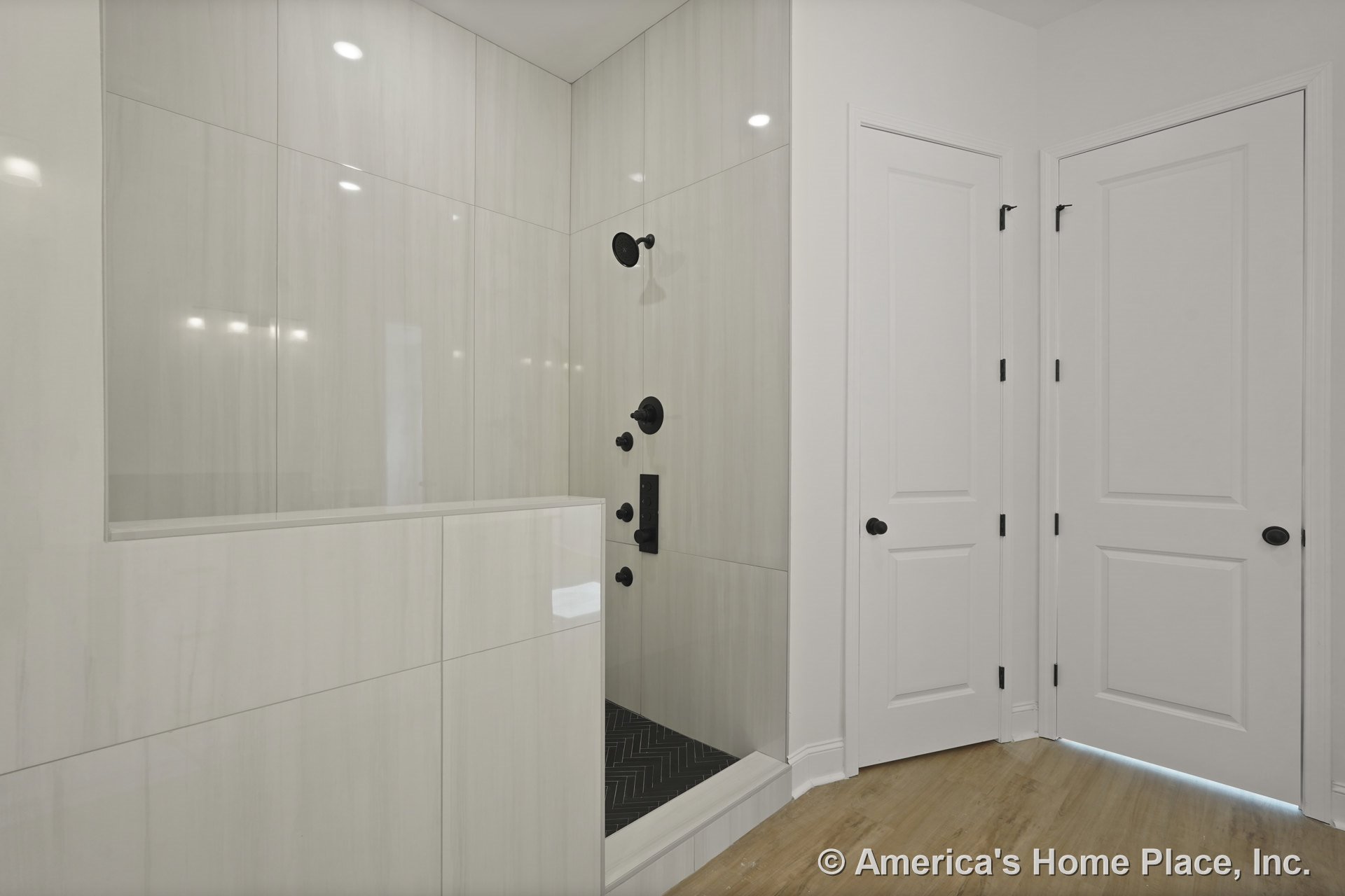 Walk-in shower with large vertical wall tiles, matte black shower fixtures, herringbone tile shower floor, white paneled doors, light wood flooring, and built-in lighting.