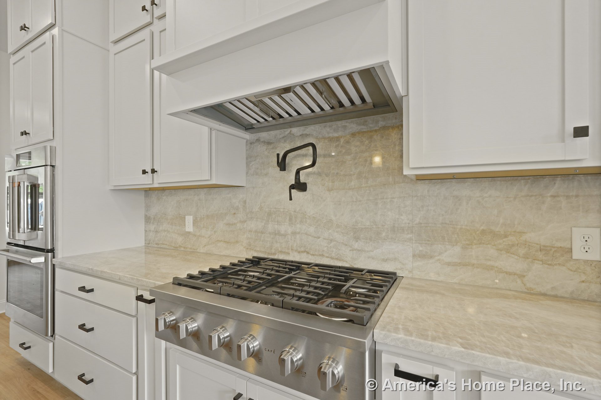 Stainless steel gas cooktop with wall-mounted pot filler set against a marble backsplash, white shaker cabinets, built-in vent hood, and stone countertops with modern cabinet