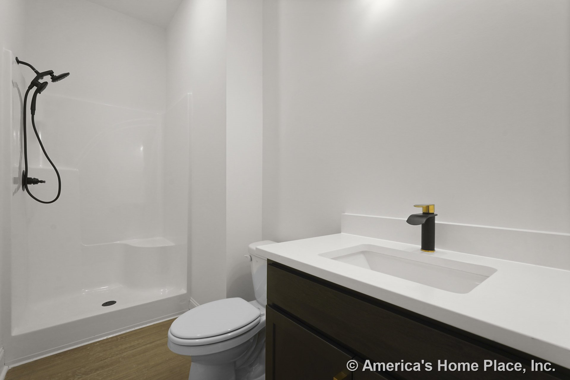 Walk-in shower with built-in seat and black handheld fixture, white solid surface vanity top, dark wood cabinetry, modern black and gold faucet, light wood flooring, white walls