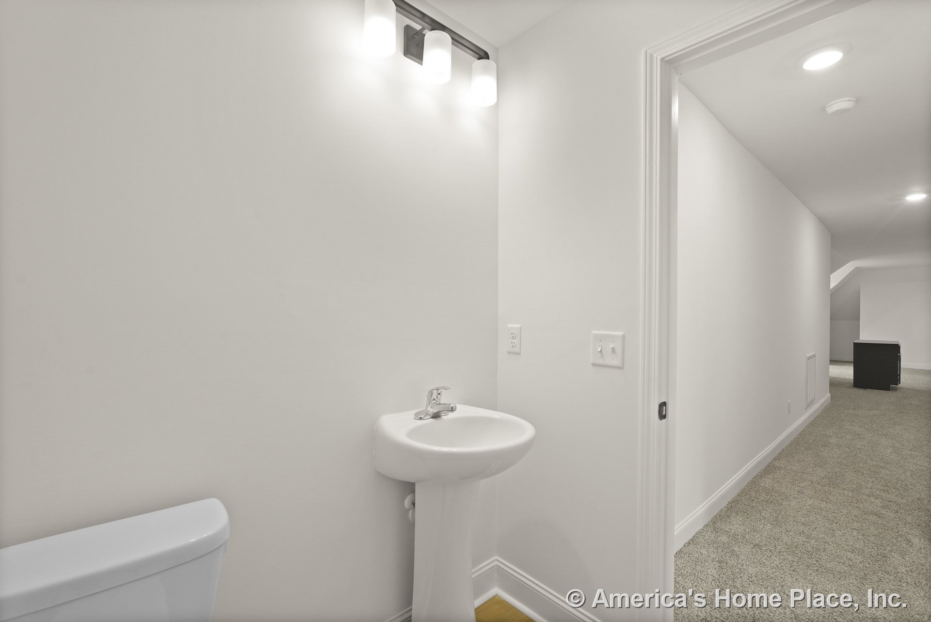 Small bathroom with pedestal sink, wall-mounted vanity light, white painted walls, recessed ceiling lights, and white baseboards; doorway opens to carpeted hallway.