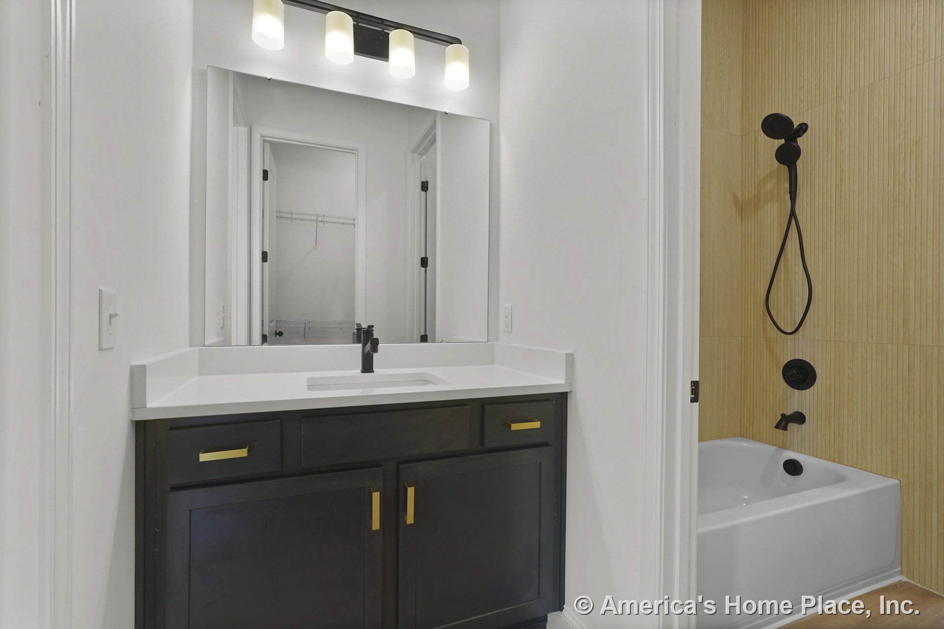 Dark wood vanity with gold hardware and white solid surface countertop, large wall mirror above, three-light vanity fixture, tub with light wood-look tile surround, modern faucet
