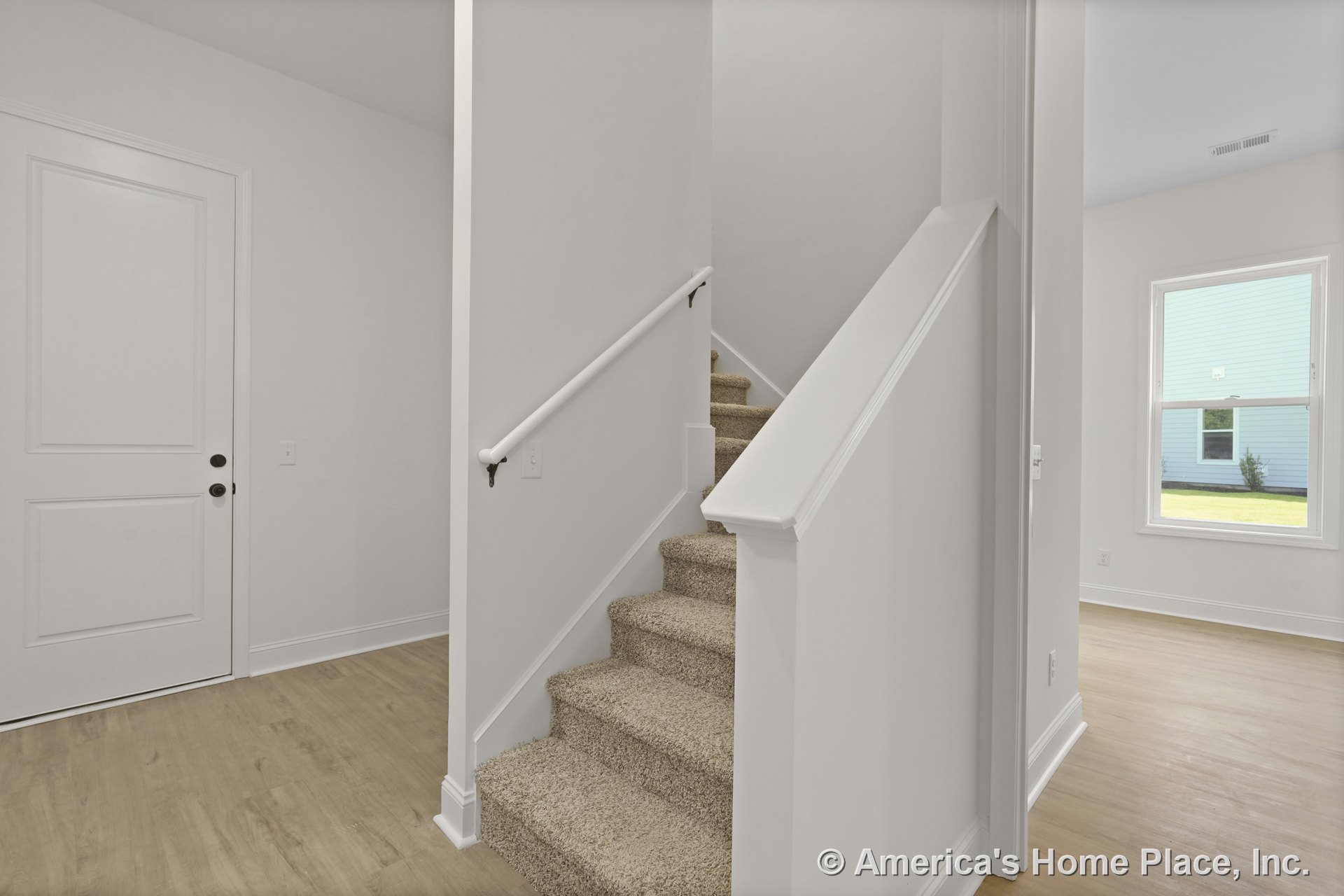 Carpeted staircase with white handrail, white paneled door, light wood flooring, white trim and baseboards, large window with white casing, neutral wall paint, and abundant natural