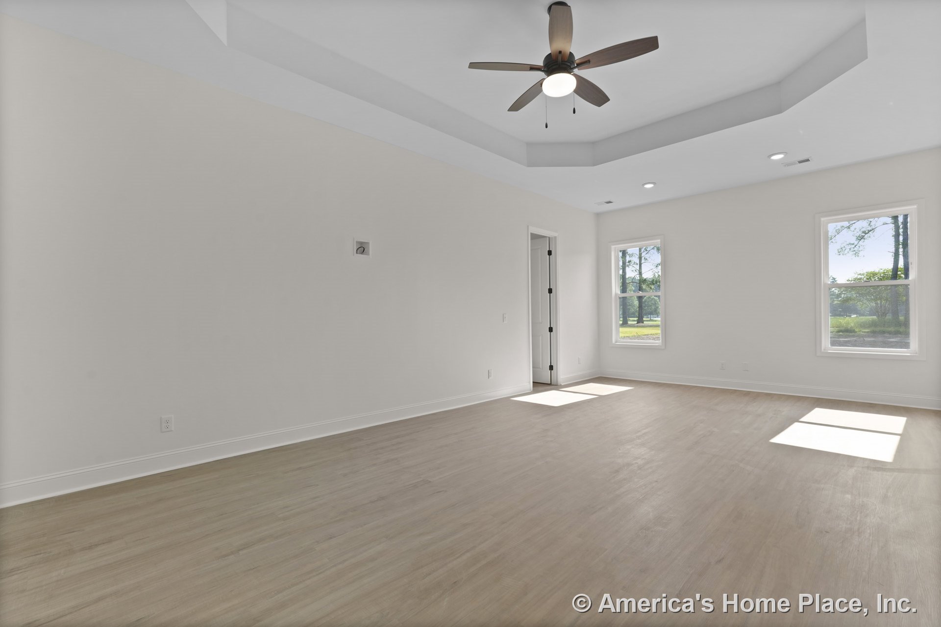 Tray ceiling with recessed lighting and ceiling fan, two large windows providing natural light, light wood plank flooring, white walls with matching trim and baseboards, interior