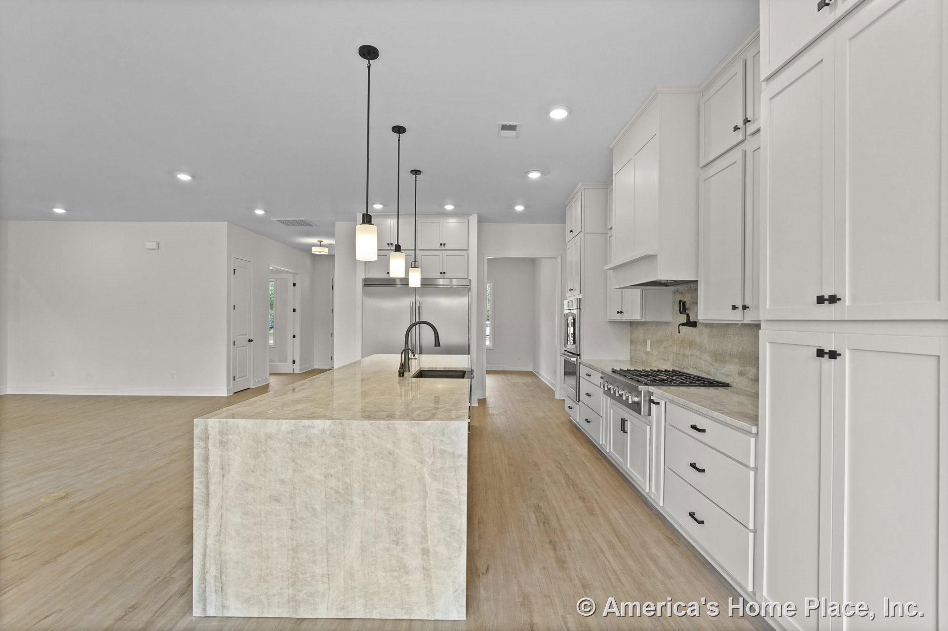 White shaker cabinetry and a large waterfall stone island with integrated pendant lighting, built-in stainless gas cooktop, double wall ovens, and refrigerator; light wood flooring