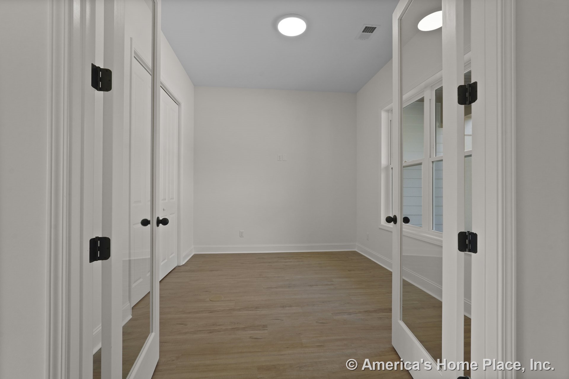 Double glass French doors open into a small interior room with wood plank flooring, large window with white trim, white walls, closet with double doors, and recessed ceiling light.