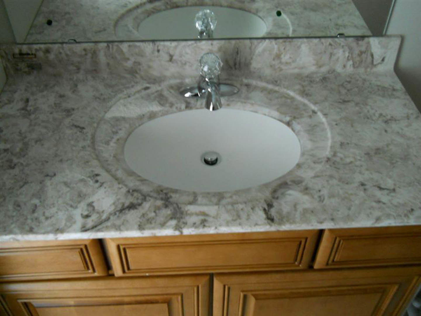 White ceramic sink with chrome faucet set in a marble countertop, rectangular mirror above, gray tile backsplash, and modern light fixture reflected in the glass.