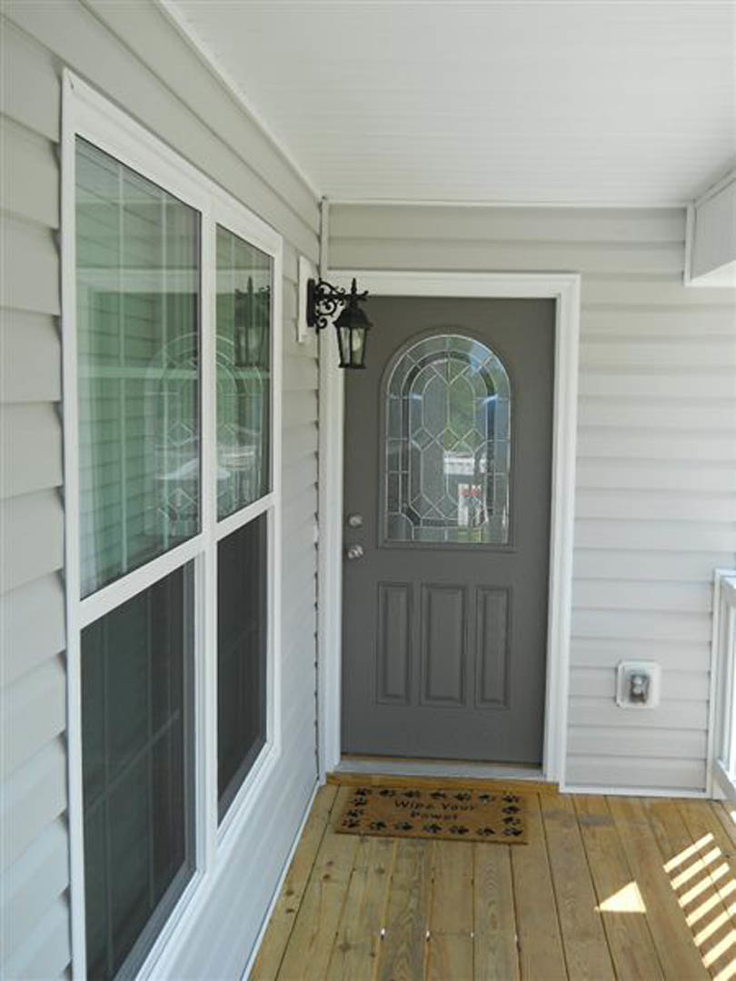 Grey door with a glass pane, wooden floor with a doormat, close-up of door handle and molding, nearby window, electrical plug, and sign.