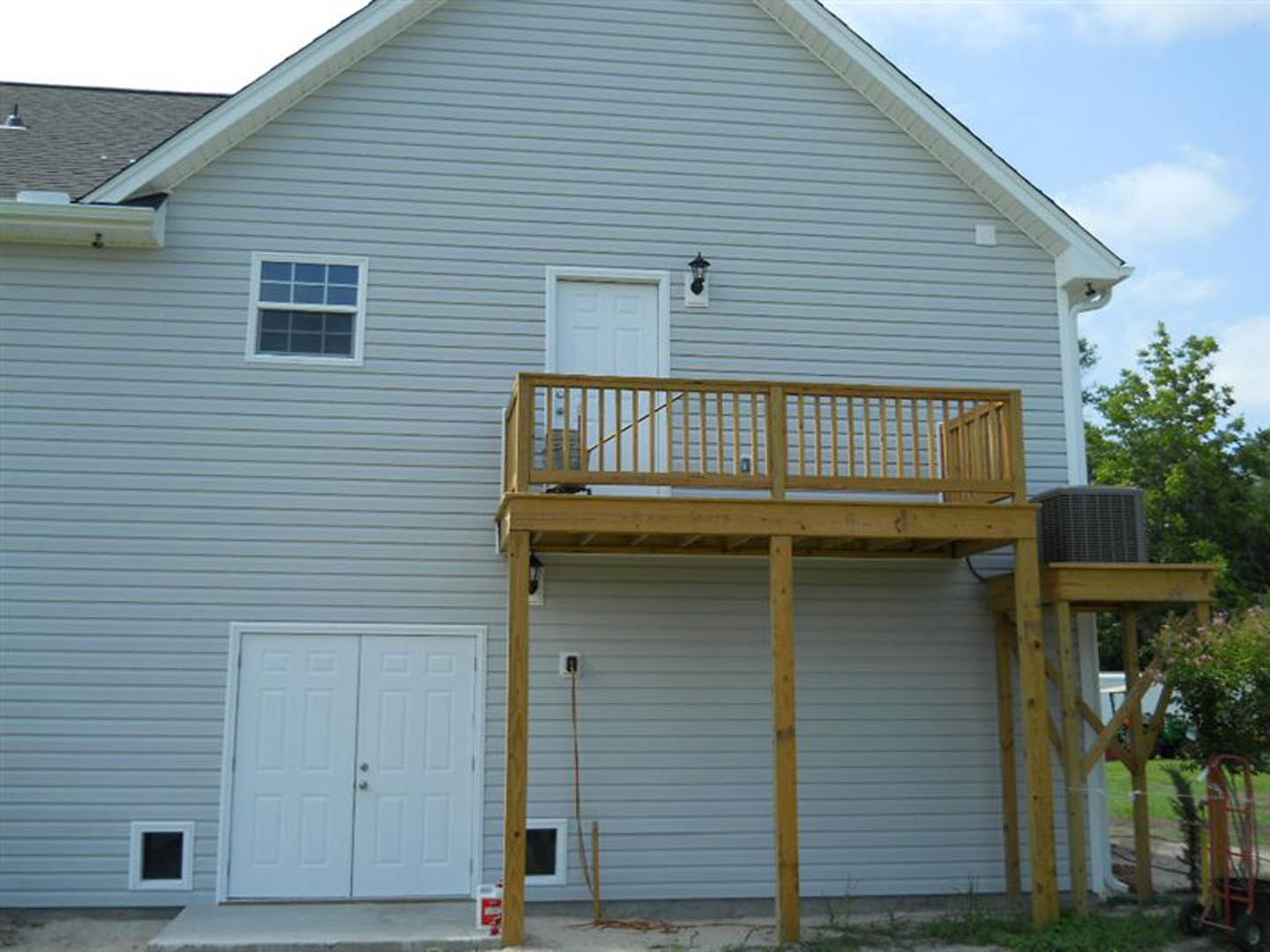 Two-story home with wood siding, upper-level balcony featuring metal railing, double glass doors opening onto a wooden deck, large windows, and covered porch beneath a gabled roof.