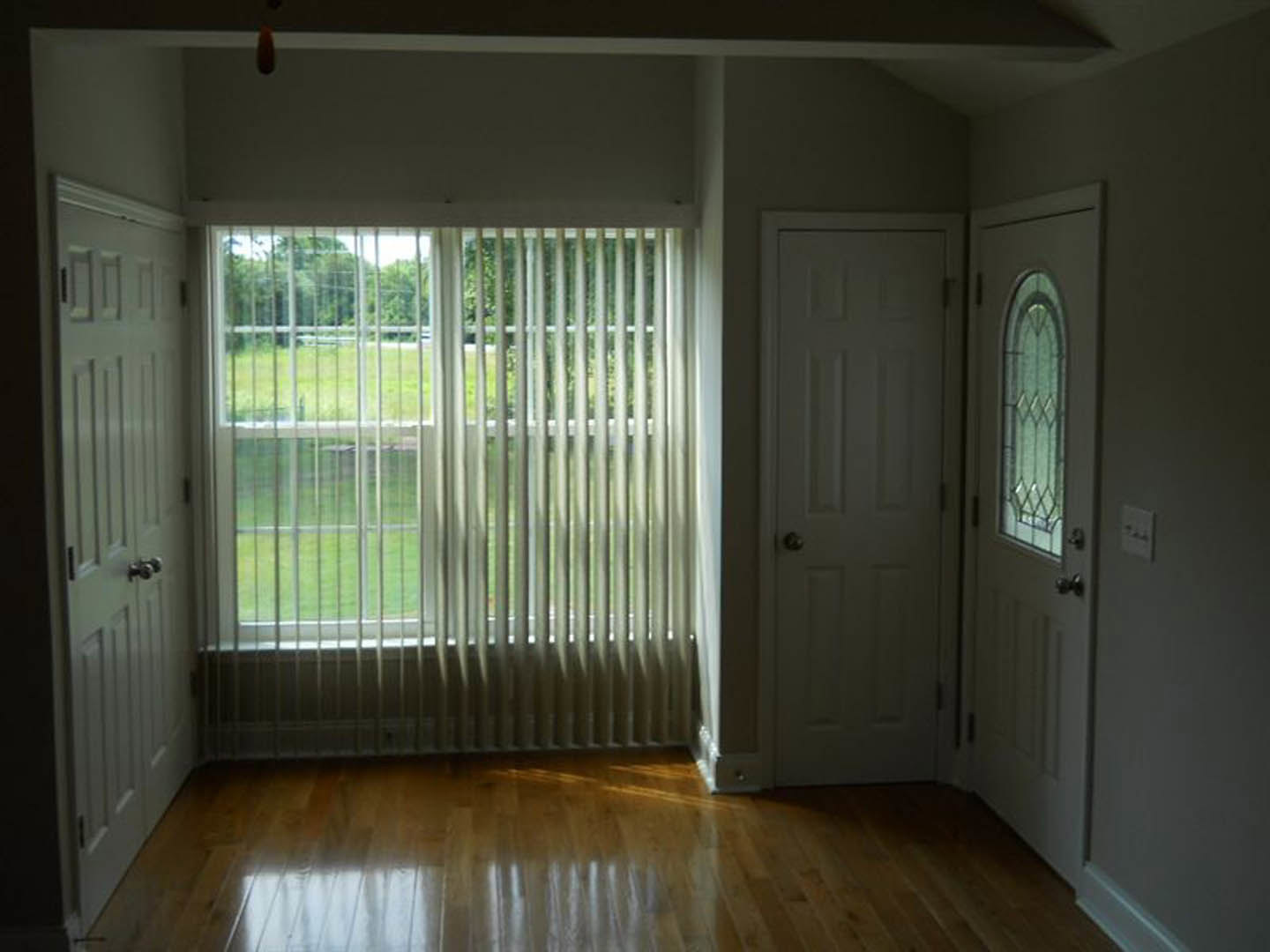 White paneled door with silver knob, window with closed blinds, light switch on pale wall, sunlight on wood laminate flooring