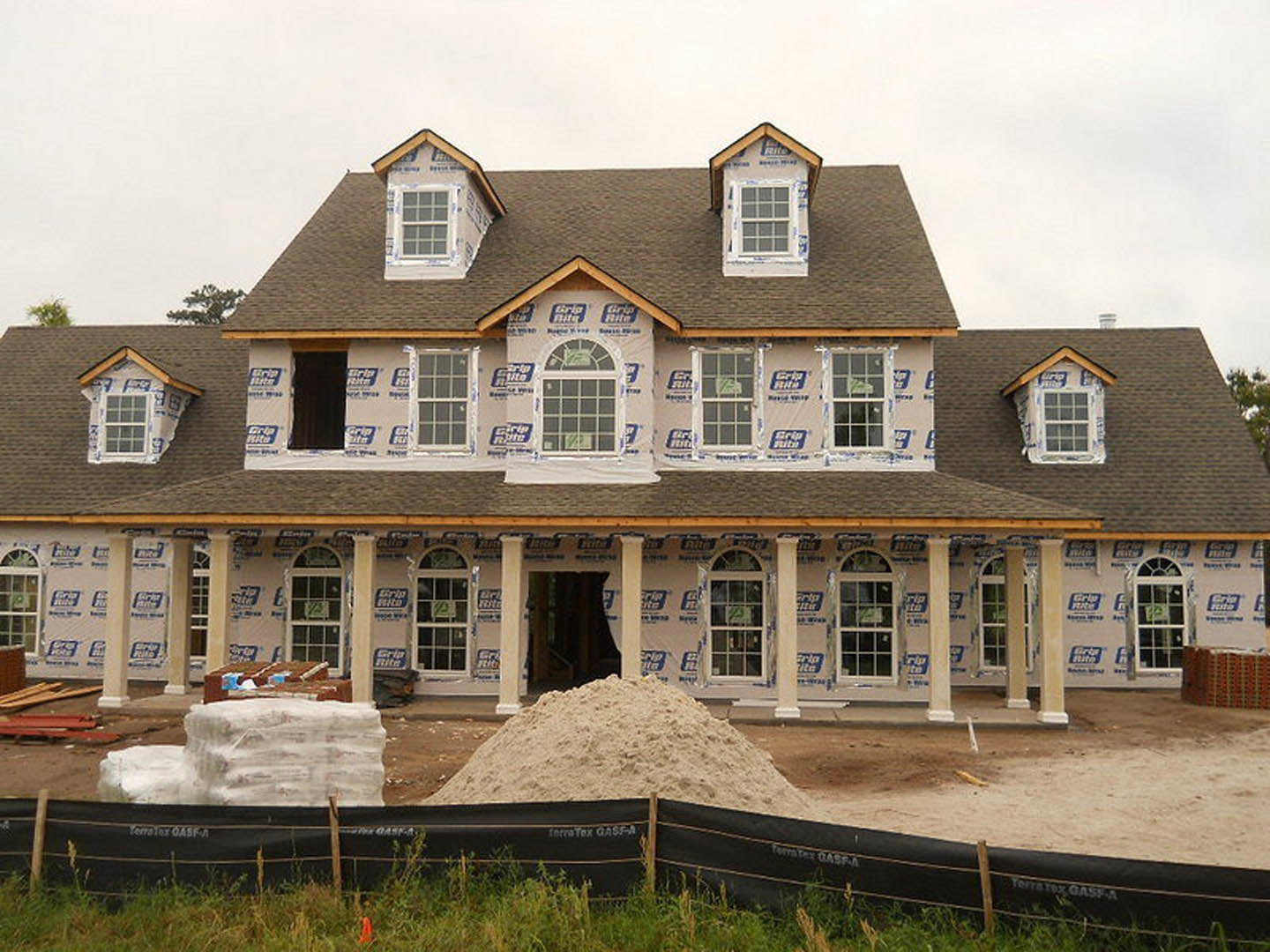 Partially built house with exposed insulation on front, sash window with multiple panes, porch area, pile of sand in a container, black tarp with white text, stack of plastic bags