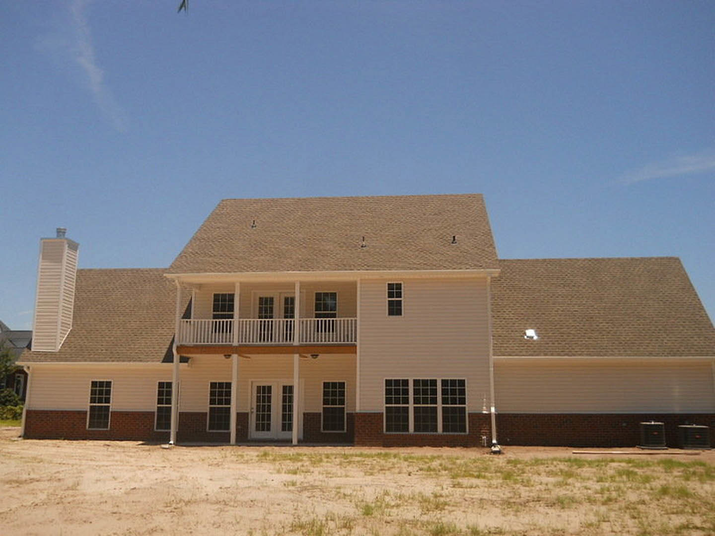 Expansive brick home with multi-pane windows, spacious covered porch, upper balcony, grassy lawn, and prominent tower structure against a cloudy sky