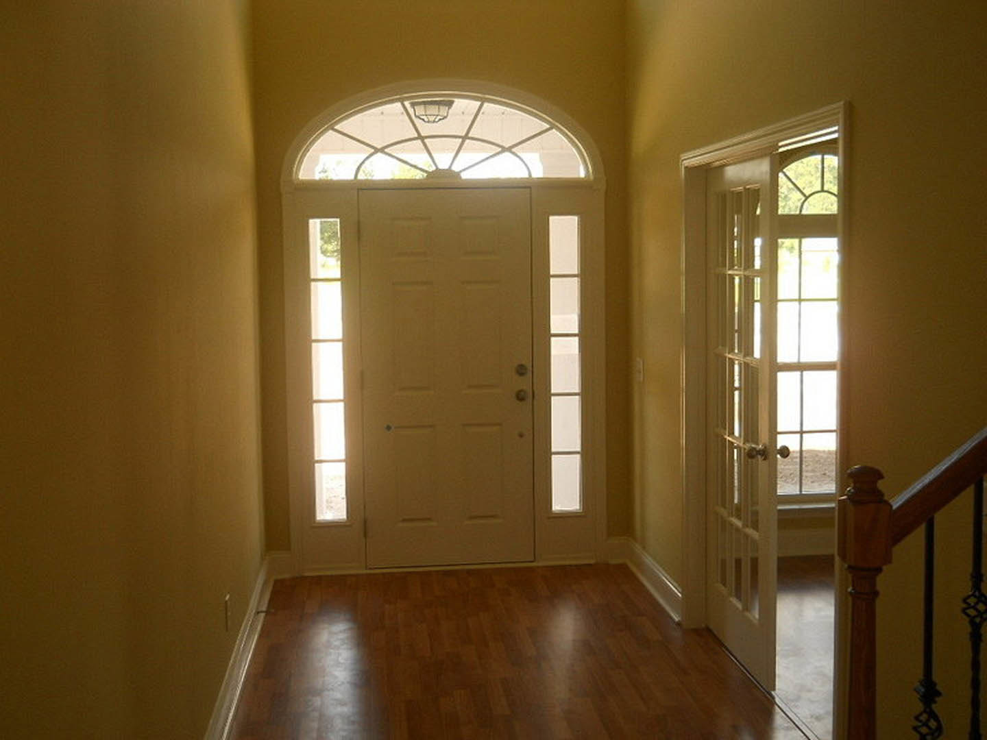 Hallway with light wood flooring, white door with glass window, wooden railing, and ceiling light fixture near a window